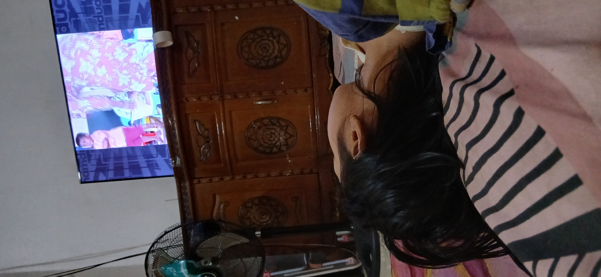 In a dimly lit room in Samarinda, Indonesia, a young person sleeps on their side, face turned away from the camera. Their dark hair is spread across a striped blanket. To their left, a black oscillating fan is visible, and beyond that, a dark wire hangs down. In the background, a wooden cabinet with decorative carvings is present. Above the cabinet, a television displays an image of a young child in a pink shirt and shorts standing next to a person wearing a pink floral patterned garment. The child is looking towards the viewer.