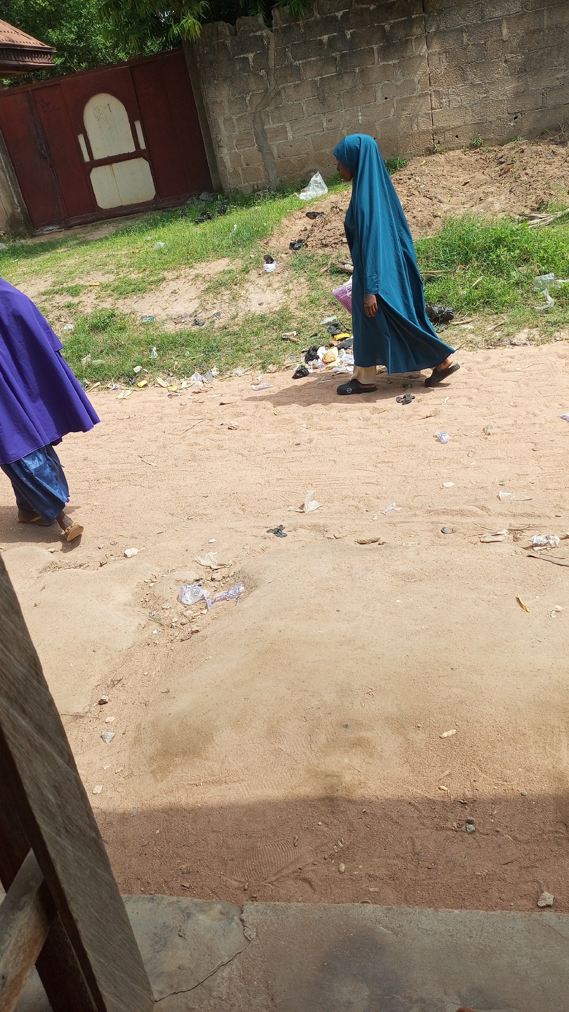 In Jalingo, Nigeria, two individuals are walking on a dirt path littered with debris. The person in the foreground, wearing a purple garment and blue trousers, is walking away from the camera. The second individual, to the right, is wearing a teal-colored hijab and a long dress, also walking away from the camera, and appears to be carrying a pink basket. The background consists of a low brick wall and a large, red metal gate with white accents. The ground is predominantly dirt with patches of green grass and scattered pieces of trash. The scene depicts a typical outdoor environment in a Nigerian city, characterized by its unpaved pathways and the presence of everyday materials.