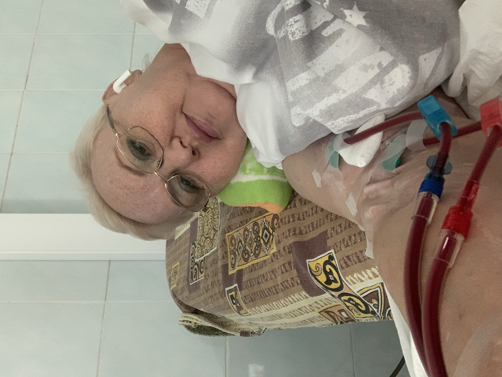 The image shows a woman with blond hair and glasses, lying on a bed with a patterned bedspread. She appears to be in a medical setting, as evidenced by the presence of medical tubing with blue and red connectors attached to her arm. She has an earpiece in her ear. The background includes tiled walls.
Given the metadata, the location is most likely in the city of Chaikovsky, Russia.