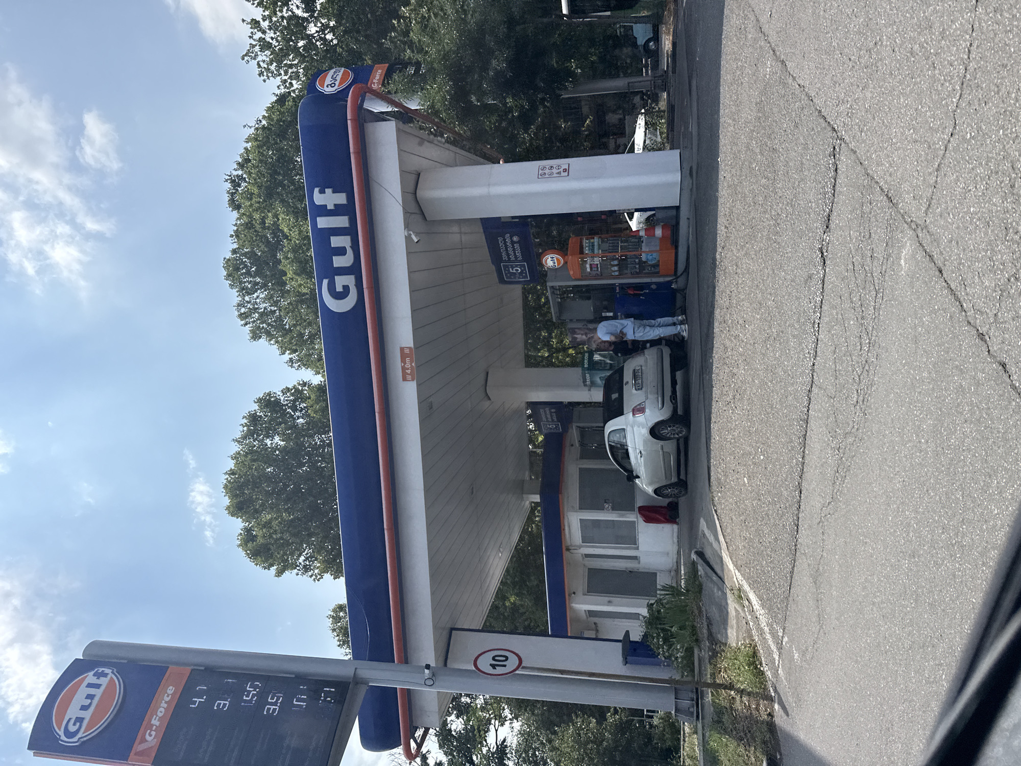 A full shot, taken outdoors on a sunny day, shows a Gulf gas station in Tbilisi, Georgia. The gas station has a blue awning with the Gulf logo prominently displayed. A white Fiat 500 is parked at one of the pumps, with a person in light blue clothing attending to it. Various signs and equipment are visible around the gas station, including a speed limit sign indicating 10, and a fuel price display. The background features lush green trees and a clear blue sky with scattered clouds. The foreground shows a cracked asphalt road.