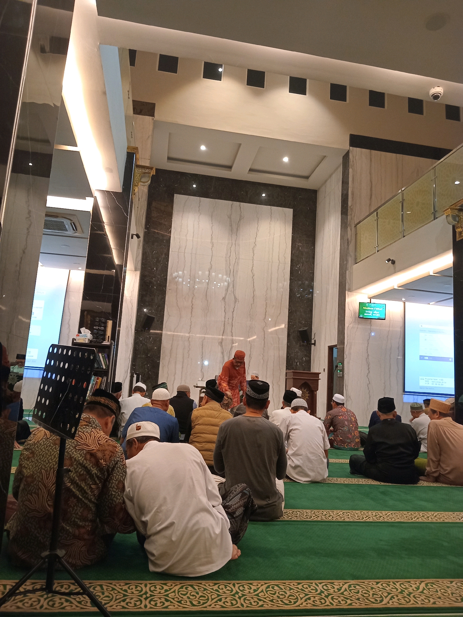 The image depicts an interior view of a mosque during a religious gathering, likely a prayer or sermon, in Randuagung, Indonesia. Several men, dressed in traditional attire including peci hats and sarongs, are seated on a green carpeted floor. One man, wearing an orange robe, stands at a podium, appearing to address the congregation. To the left, a music stand is visible, and in the background, large projection screens display text and images. The mosque's architecture features marble walls, modern lighting, and security cameras, suggesting a contemporary place of worship. The overall atmosphere appears to be solemn and respectful.