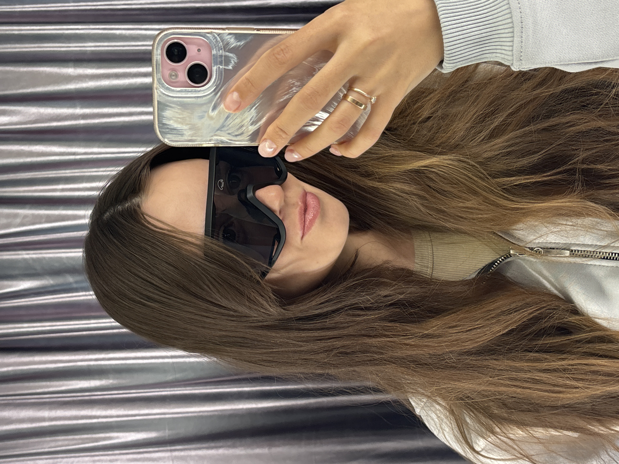 A young woman is taking a selfie with her smartphone in what appears to be an indoor setting. She is wearing sunglasses and holding the phone above her head. She has long brown hair. She is wearing a light-colored sweater with a zipper. She has a ring on her finger. The background is a shiny, metallic-looking surface.
There is no information in the image that places it in the city of Krasnoyarsk, Russia.