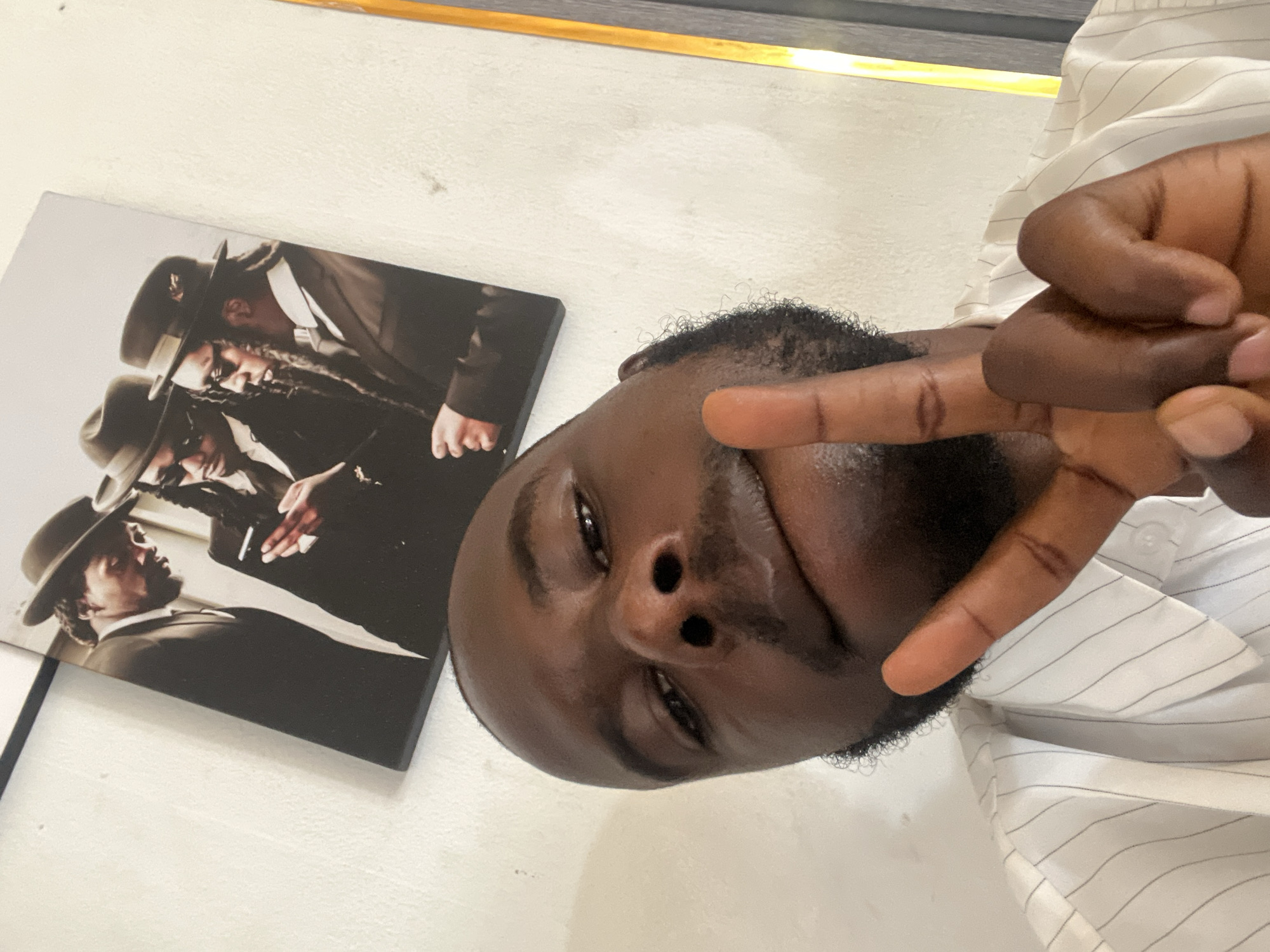 A close-up image shows a man with dark skin and a short beard, making a peace sign with his right hand. He is wearing a white shirt with thin gray stripes. Next to him is a photograph of three men in black suits and fedora hats. The background is a plain white wall. There is no indication of the city of Gura Top, Nigeria.