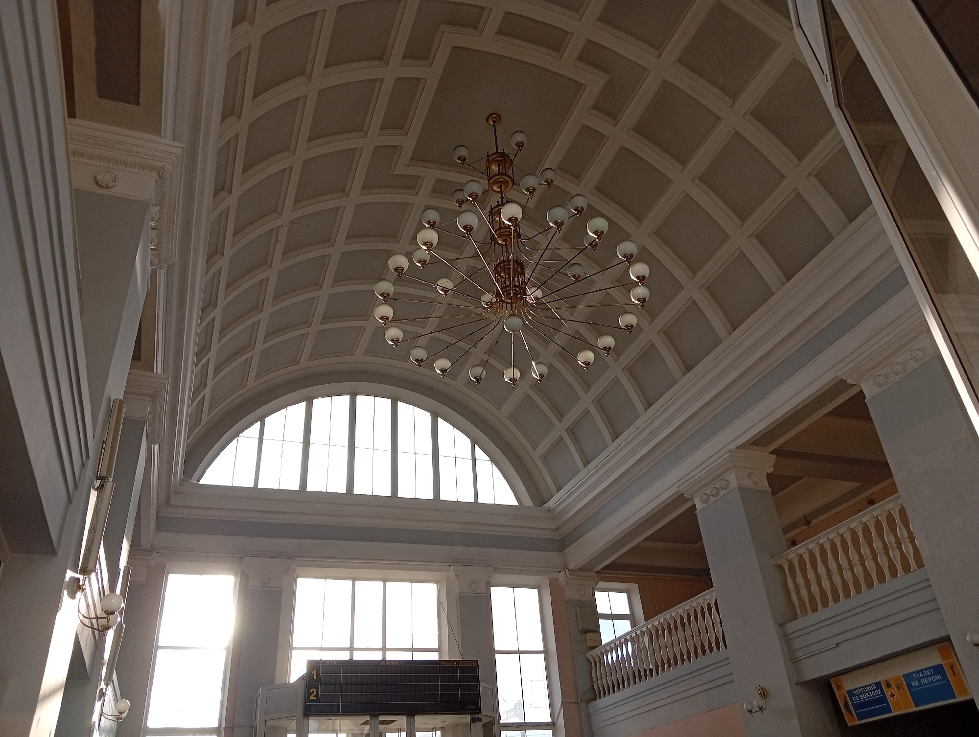 The image depicts the interior of a building, likely a train station, in Poltava, Ukraine. The architecture features high ceilings with coffered patterns and large arched windows allowing daylight to illuminate the space. A prominent chandelier hangs centrally from the ceiling. Below, an electronic timetable displays train information. To the right, a balcony with balustrades overlooks the main hall, and signage indicates directions, possibly to restrooms. The overall environment suggests a public transportation hub designed with classical architectural elements. There are no people visible in the image.