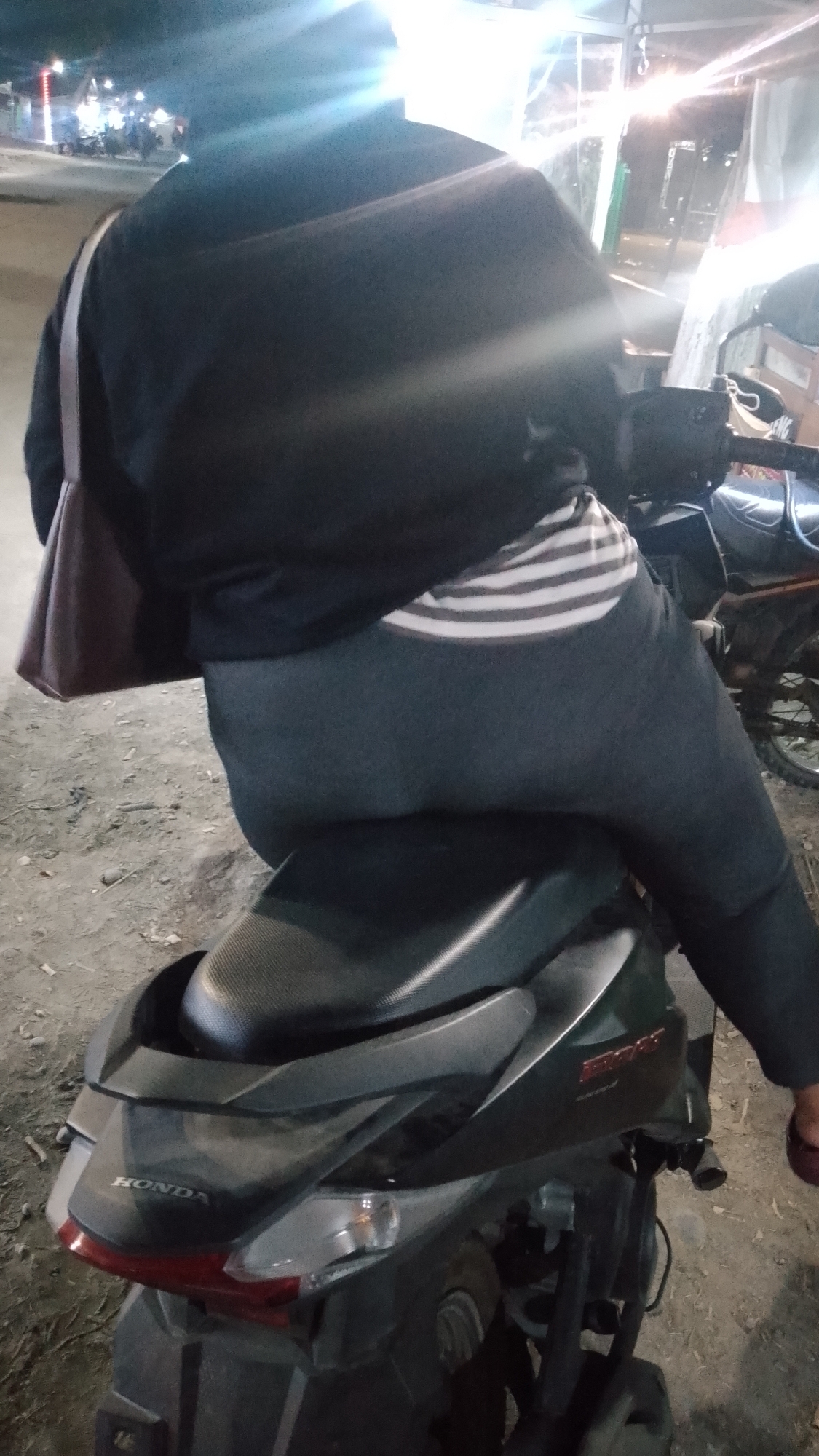 A person is sitting on a dark grey Honda scooter at night on a dirt road in Karanglangu, Indonesia. The person is wearing a black jacket, grey pants, and a striped shirt. A brown shoulder bag is slung across their back. The scooter has a black seat and a red taillight. In the background, there are streetlights and what appear to be market stalls with lights.