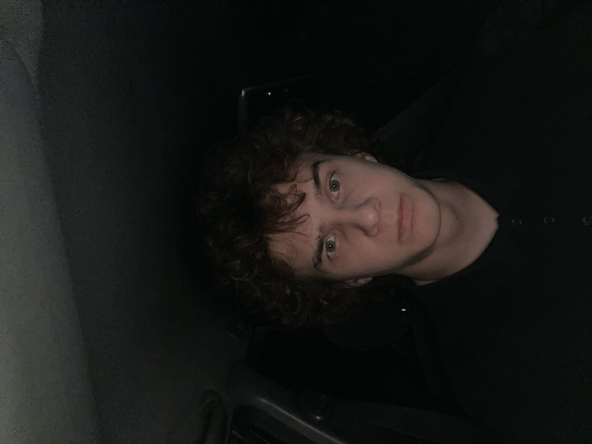 A young male with curly brown hair, wearing a black shirt, is pictured in a dimly lit environment. The lighting suggests the photo was taken at night, possibly inside a vehicle given the visible interior elements and the darkness outside. His expression is neutral. No specific location in Krasnoyarsk, Russia, can be determined from the image.