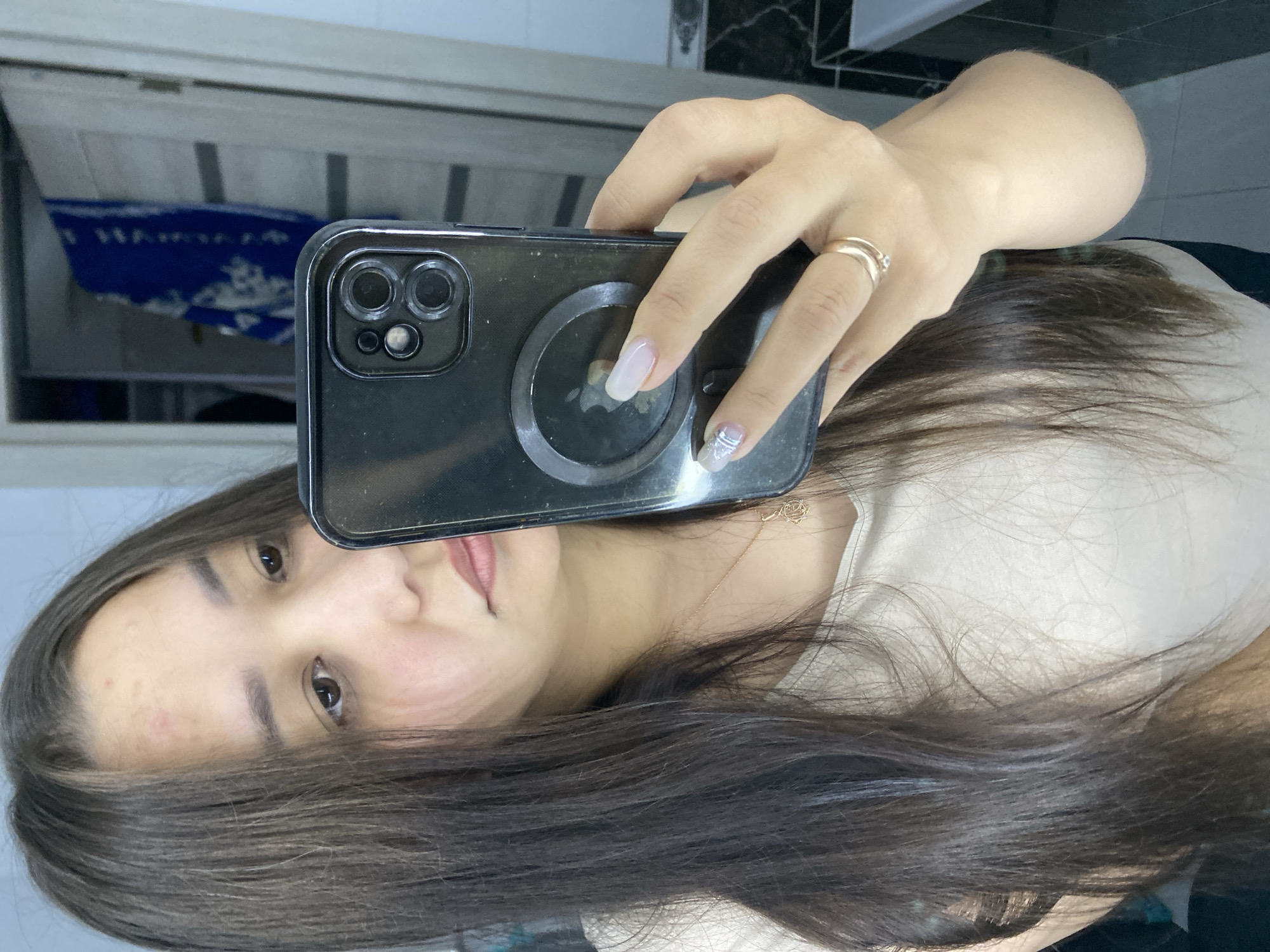 A young woman is taking a mirror selfie in what appears to be a bathroom. She has long, dark brown hair and is wearing a light-colored t-shirt. She is holding a black smartphone with a clear case, which has a magnetic ring on the back. She is wearing a gold ring on her left ring finger and has manicured nails. The background shows a doorway with a blue towel hanging on it, and a tiled wall. The lighting is bright and natural. It is not possible to determine the specific location within the city of Ufa, Russia.