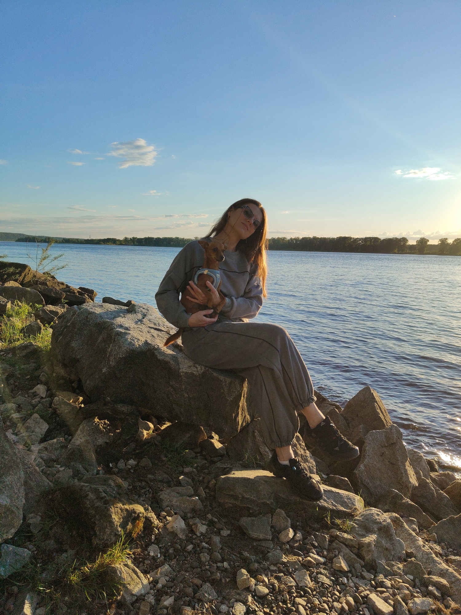 A young woman is seated on large rocks by the edge of a body of water, holding a small, light brown dog. She has long brown hair, wears sunglasses, and is dressed in a casual grey sweatsuit with black sneakers. The dog, possibly a chihuahua or similar small breed, is wearing a light blue harness and appears comfortable in her arms.
The setting is a natural outdoor environment, likely a riverbank or lake shore. The water is calm, reflecting the sky, and a distant treeline is visible on the opposite bank. The rocky foreground includes some sparse green grass and foliage. The sky above is clear and blue with a few scattered white clouds.
The image captures the golden hour, indicating late afternoon or early evening, as warm sunlight casts long shadows and creates a lens flare effect on the right side of the frame. The woman appears relaxed, looking towards the viewer with a slight smile. There is no visible text in the image.