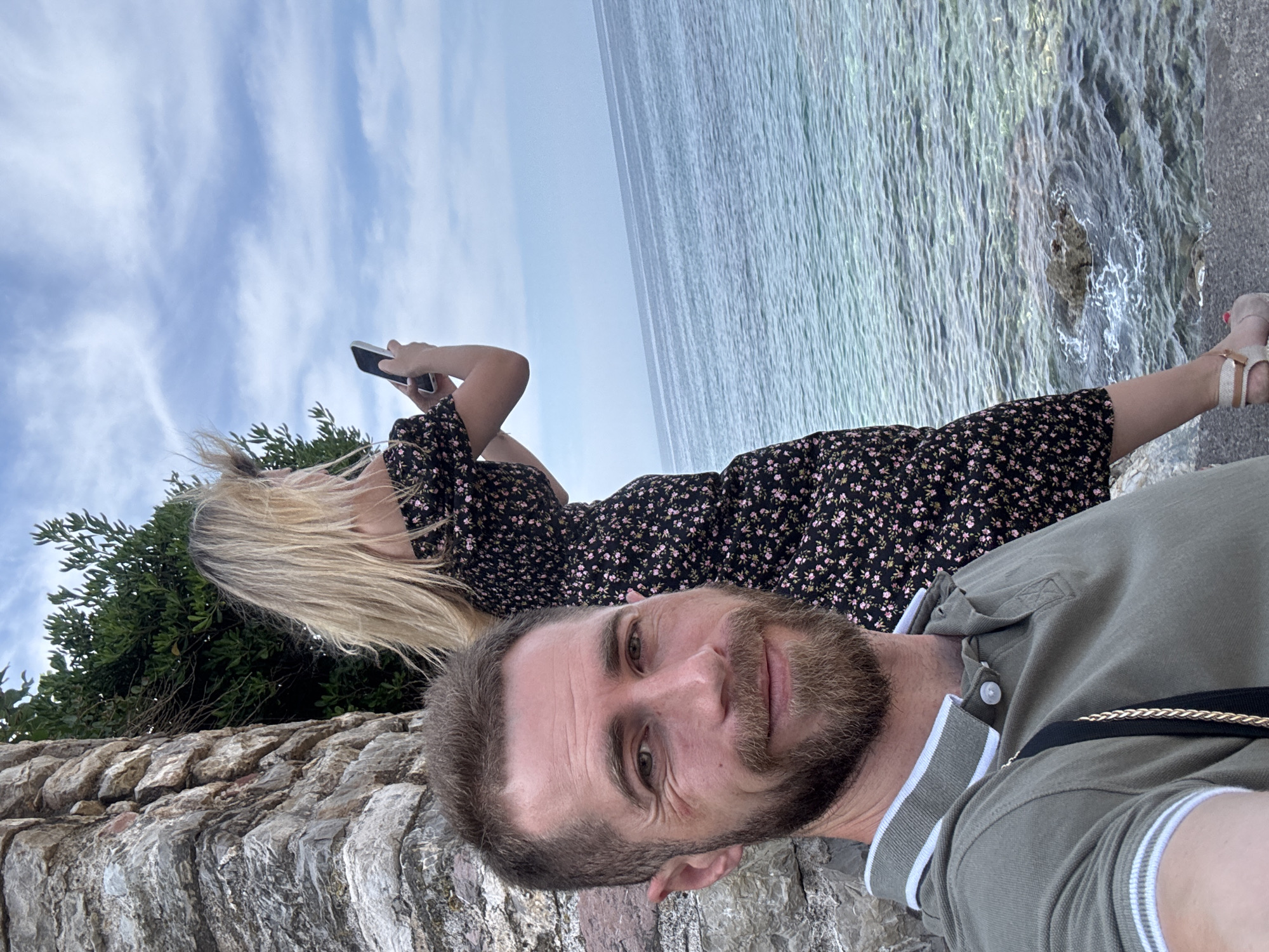 A man with a beard and a light olive-colored polo shirt is taking a selfie outdoors, leaning against a textured stone wall. Behind him, a blonde woman wearing a black floral dress is standing, facing away from the camera, and holding a smartphone to take a picture. They are positioned near the sea, with clear blue water and a rocky shoreline visible. The sky is partly cloudy. The man's gaze is directed at the camera, and he has a slight smile. The woman appears to be focused on her phone. The scene suggests a casual outing or vacation by the coast, possibly in Budva, Montenegro, given the rocky coastline and clear water often found in the region.