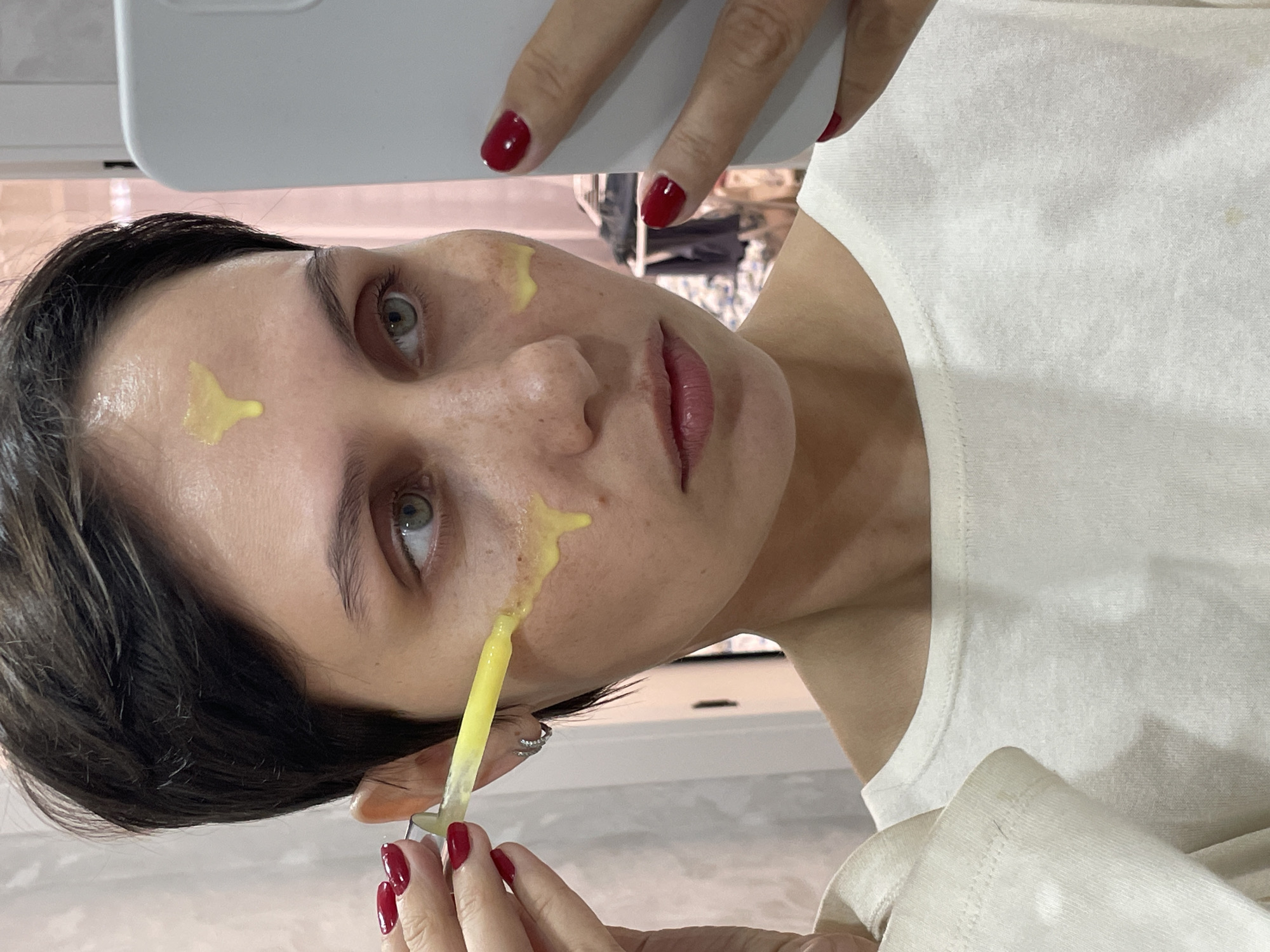 A woman with short, dark hair and fair skin is holding a white smartphone in her left hand, taking a selfie. She is wearing a white top. Her fingernails are painted red. She has a yellow substance applied to her face in several spots. She is applying more of the substance from a tube or applicator in her right hand. The background shows a blurred indoor setting, likely a bathroom or bedroom, but it is not possible to determine the exact location in Izhevsk, Russia, from this image.