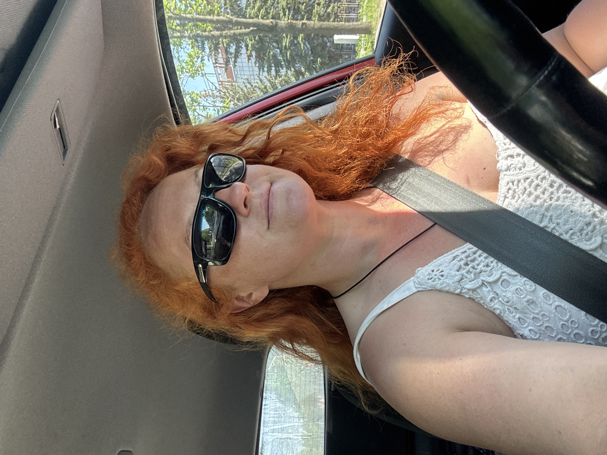 A woman with red curly hair and wearing sunglasses is inside a vehicle, seemingly driving. She is wearing a white, crocheted top and a seatbelt is fastened across her body. The steering wheel is visible in the foreground. Outside the car window, trees and possibly a building with scaffolding are visible. There is no definitive evidence to confirm the location is Chaikovsky, Russia.
