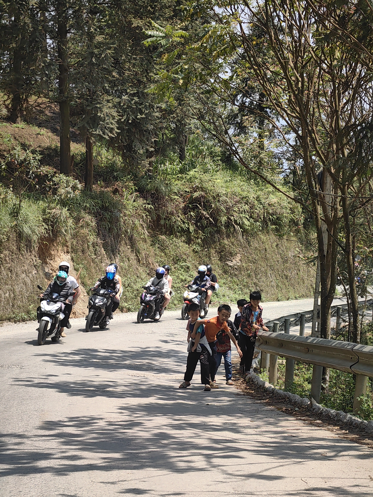 The Yên Minh Commune, Vietnam scene shows a winding road with a group of children walking alongside a metal guardrail. A line of motorcyclists, all wearing helmets, is traveling along the road toward the children. The surrounding environment includes trees and vegetation covering the hillside. The road surface is paved, and shadows from the trees are visible on the road.