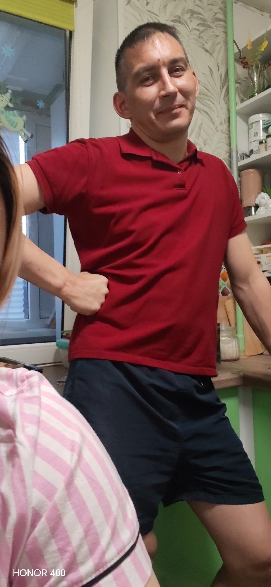 The image depicts a man wearing a red polo shirt and navy blue shorts. He has short, dark hair and is standing with his right hand on his hip. He appears to be indoors, possibly in a kitchen. The background includes a window with decorations and a shelf with various items on it. Another person is partially visible in the bottom left corner, wearing pink and white striped clothing. The text "HONOR 400" is visible in the bottom left corner. There is no verifiable information to support the city location mentioned.
