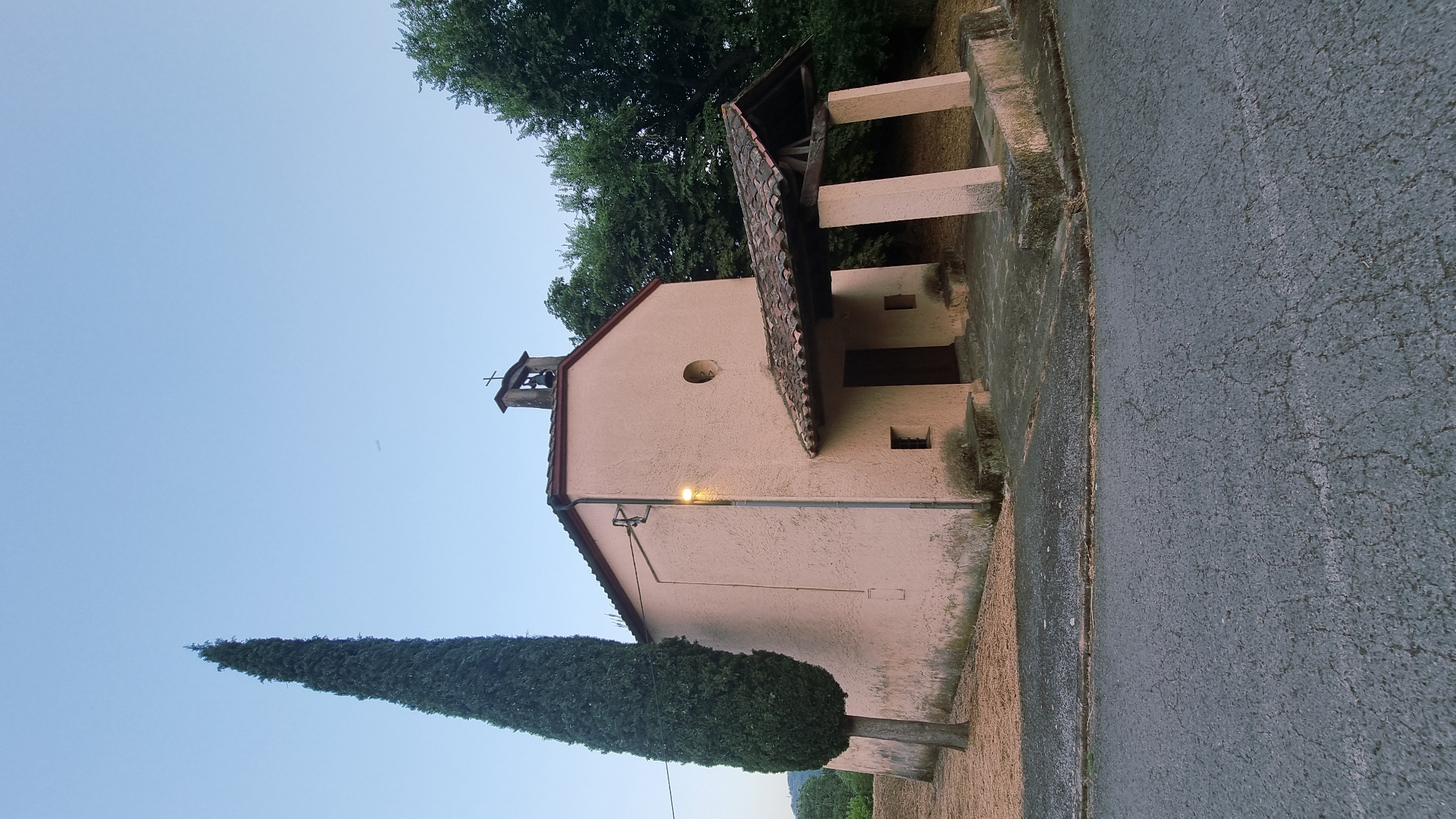 The image shows a small, pink stucco church with a red tile roof, located in La Vall de Bianya, Spain. A tall, slender cypress tree stands to the left of the church, and a paved road is in the foreground. The sky is clear and pale blue, with a faint contrail from an airplane visible. The church has a bell tower with a cross, a covered porch with wooden beams and pink columns, and a small, round window above the entrance. The area surrounding the church appears to be rural, with trees in the background. The lighting suggests it is either early morning or late evening.