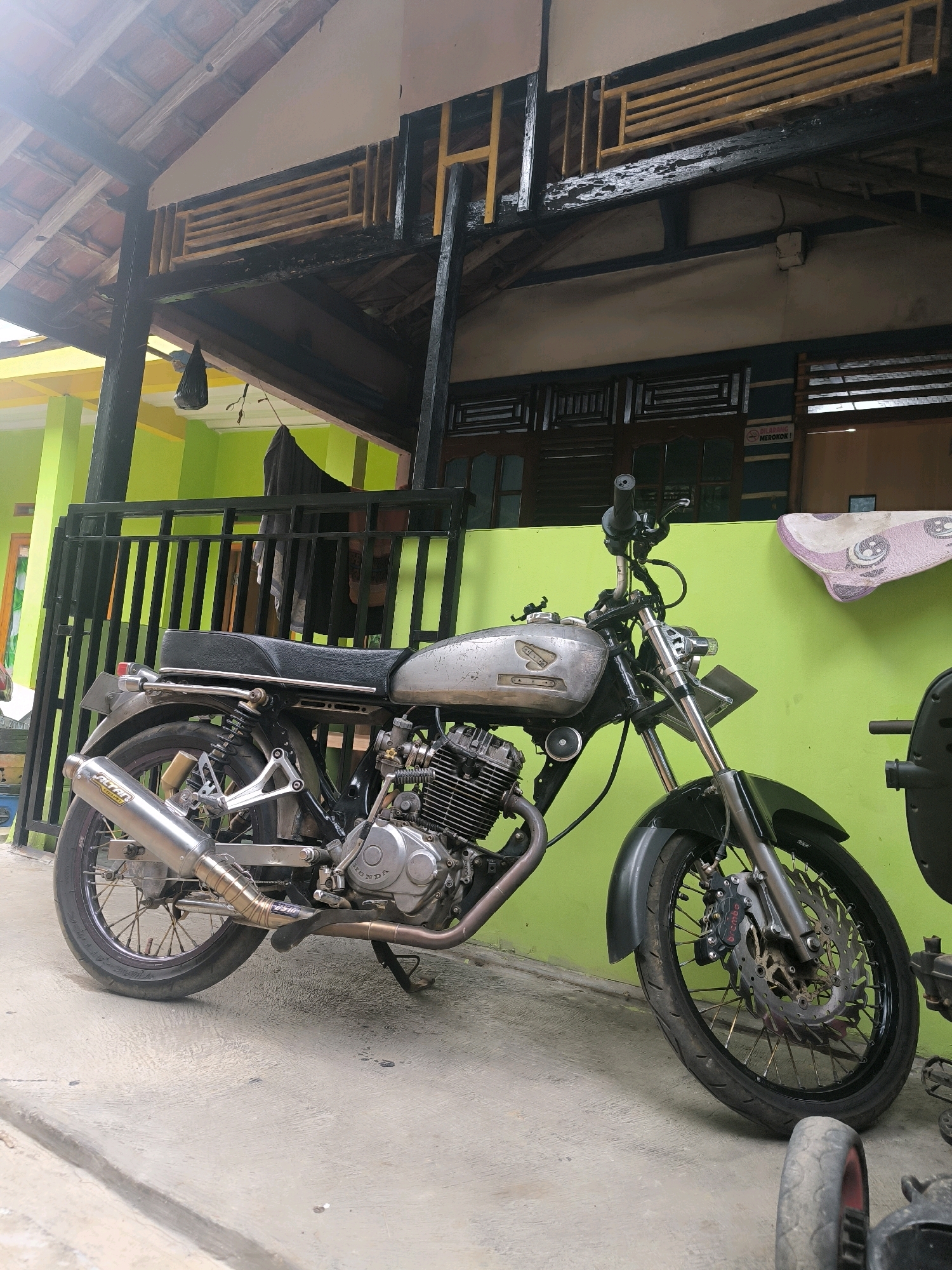 A silver motorcycle is parked on a concrete ground. The motorcycle has a black seat and a metallic exhaust pipe. The motorcycle is positioned in front of a bright green wall, with a black metal fence behind it. The scene appears to be outdoors, possibly in a residential area, given the presence of a roof with terracotta tiles overhead. The overall environment suggests a warm, possibly humid climate. The image does not depict any people or actions. The location is specified as Cilengkrang, Indonesia.