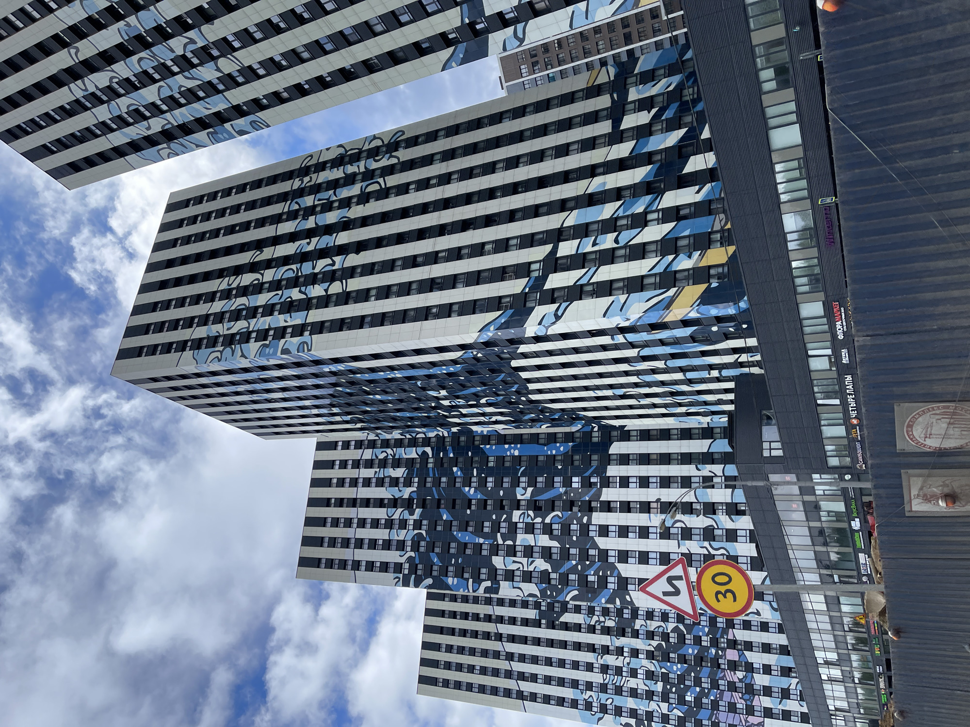 The image depicts a view of a modern architectural complex in Moscow, Russia, characterized by its tall, high-rise buildings with horizontally striped facades. The buildings feature a distinctive blue and white artistic pattern superimposed on the striped structure. The sky is partially visible, showcasing white clouds against a blue background between the buildings. A yellow and red circular traffic sign with the number "30" is visible, along with another sign showing a curving road. Part of the street level is seen on the right, with visible storefronts. The image is taken from a low angle.