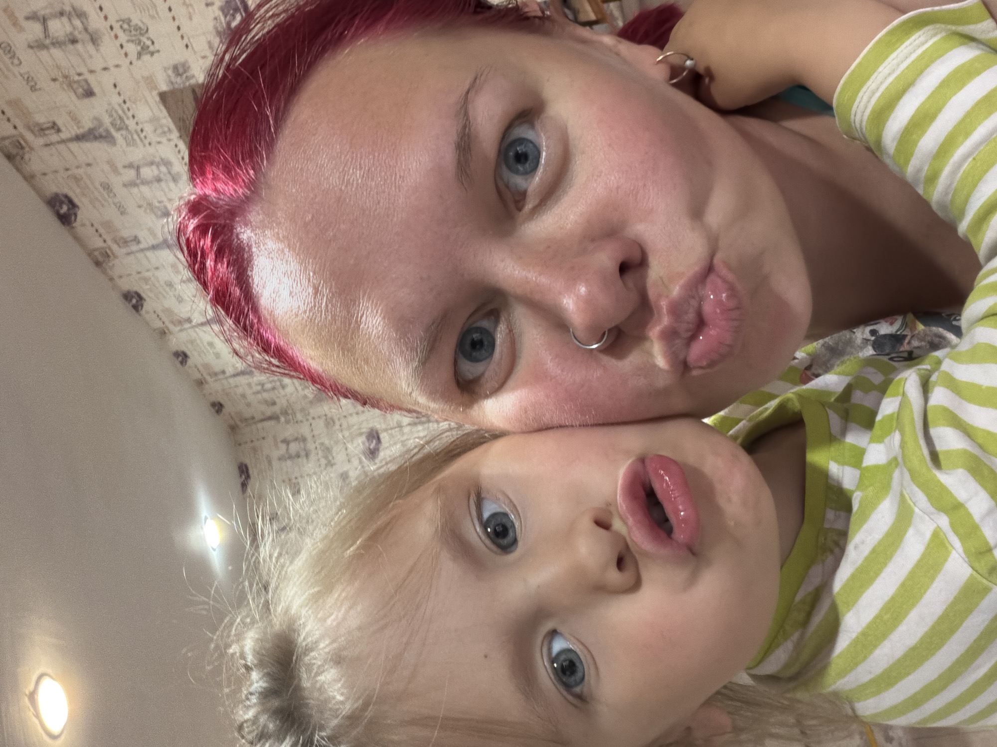 The image captures a close-up selfie of two individuals in an unknown indoor location. On the right, an adult female with red hair, light blue eyes, a silver hoop nose ring on her left nostril, and small hoop earrings, directly faces the camera with her lips pursed. Her skin exhibits a reddish tone. Partially visible on the left, a young blonde-haired child with light blue eyes leans into the adult, also directly facing the camera with pursed lips, mimicking the adult's expression. The child is wearing a green and white striped long-sleeve top. The child's left arm is visible around the adult's shoulders/back. The background consists of a light-colored wall adorned with a patterned wallpaper featuring sketches of landmarks and text such as "POST CARD." Overhead, multiple recessed ceiling lights provide illumination.