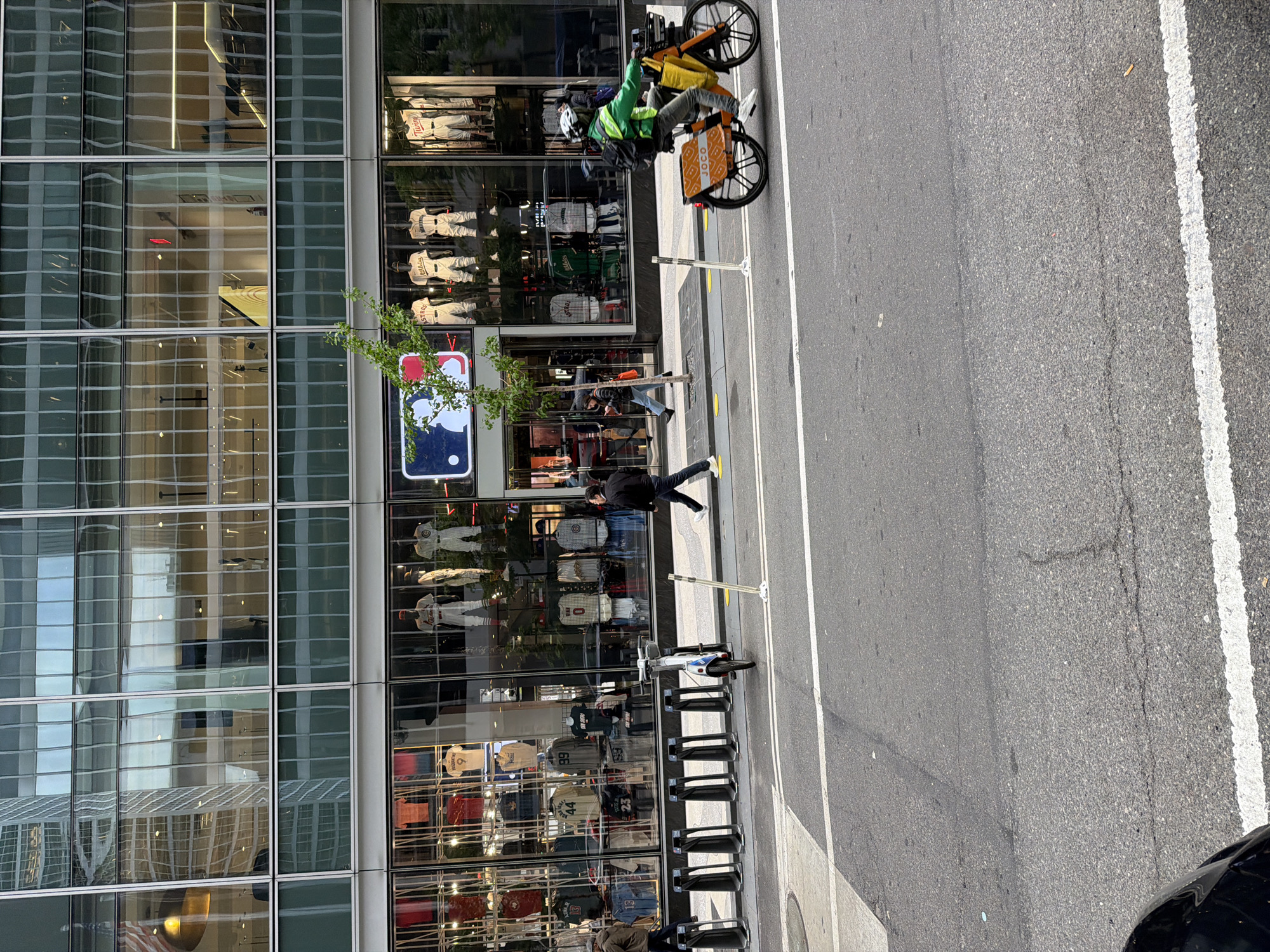An outdoor urban scene captures a segment of a city street and a modern building, set in New York, United States of America. The image is oriented vertically with the building on the left and the street on the right.
The prominent feature on the left is a multi-story building with a facade composed mostly of large, reflective glass windows, showcasing reflections of surrounding structures and the sky. The ground floor houses a store, indicated by display windows filled with various sports jerseys and uniforms on mannequins or hangers. Visible team names on jerseys include "Twins" and "Astros," along with numbers like "99" and "44." A large, illuminated sign featuring the distinctive Major League Baseball (MLB) logo is visible within one of these windows, partially obscured by a small green tree.
Adjacent to the building is a wide concrete sidewalk. A person in a black jacket and dark pants walks along the sidewalk towards the right. A row of empty black metal bicycle racks stretches along the sidewalk, and a white and blue electric scooter is parked nearby.
To the right of the sidewalk is an asphalt street with white painted lines marking lanes. A delivery person on a bright orange cargo bicycle is a central subject in the upper part of the image. They wear a bright green jacket or vest and a white helmet, appearing to be in motion. The bicycle features a large front basket with a yellow bag and a distinctive orange cargo box at the rear with the text "JOCO" clearly visible. The overall impression is a bustling daytime street in a major city.