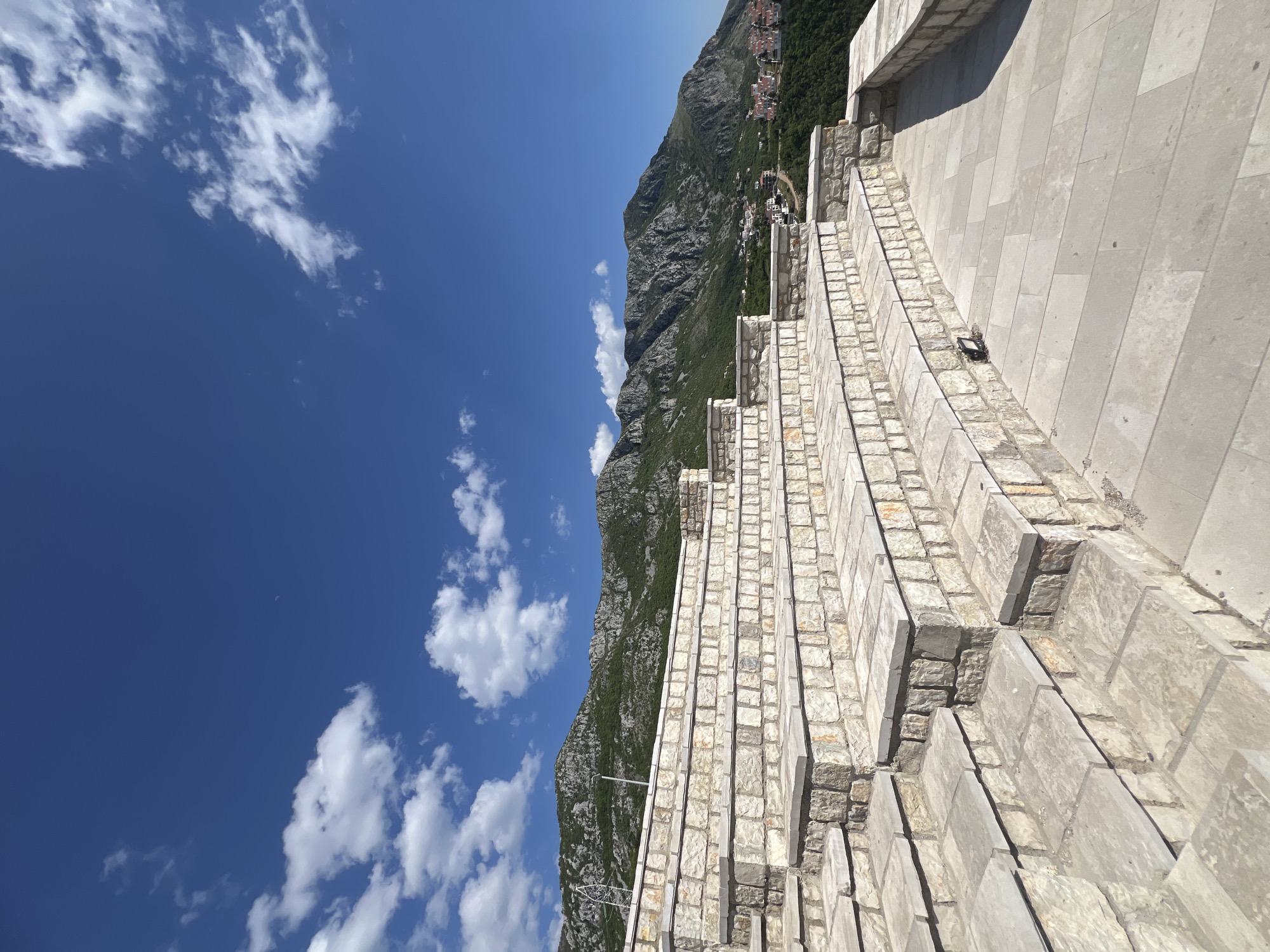 An outdoor, rotated image captures a vast landscape featuring a prominent multi-tiered stone structure in the foreground, leading up towards a majestic mountain range under a bright blue sky.
The main subject is a wide, terraced staircase or stepped wall made of light-colored, rectangular stone blocks. The steps vary in width and depth, creating a dynamic architectural element. Sunlight casts clear shadows on the right side of the steps, indicating the sun is overhead or to the left. The stone work is detailed, showing individual blocks and mortar lines. Further up the structure, a smooth, lighter-colored retaining wall is visible.
Beyond the stone structure, a large, rugged mountain dominates the midground and background. The mountain is mostly covered in dark green vegetation, suggesting dense trees and bushes, with exposed rocky patches revealing grey stone. In the distance, nestled on the slopes of the mountain, are clusters of small buildings and houses, some with distinctive red or terracotta roofs, hinting at a small town or settlement.
The sky, occupying a significant portion of the image, is a vibrant, clear blue with scattered white, fluffy cumulus clouds. These clouds are irregularly shaped and spread across the expanse, adding depth to the otherwise clear sky.
The scene depicts a sunny day, likely during the morning or afternoon, given the strong illumination and defined shadows. The overall impression is one of natural beauty combined with significant human-made construction, possibly a monument or a pathway offering scenic views. Based on the provided context, the location is Sveti Stefan, Montenegro, which is consistent with the mountainous coastal landscape and stone architecture. No visible text is present in the image.