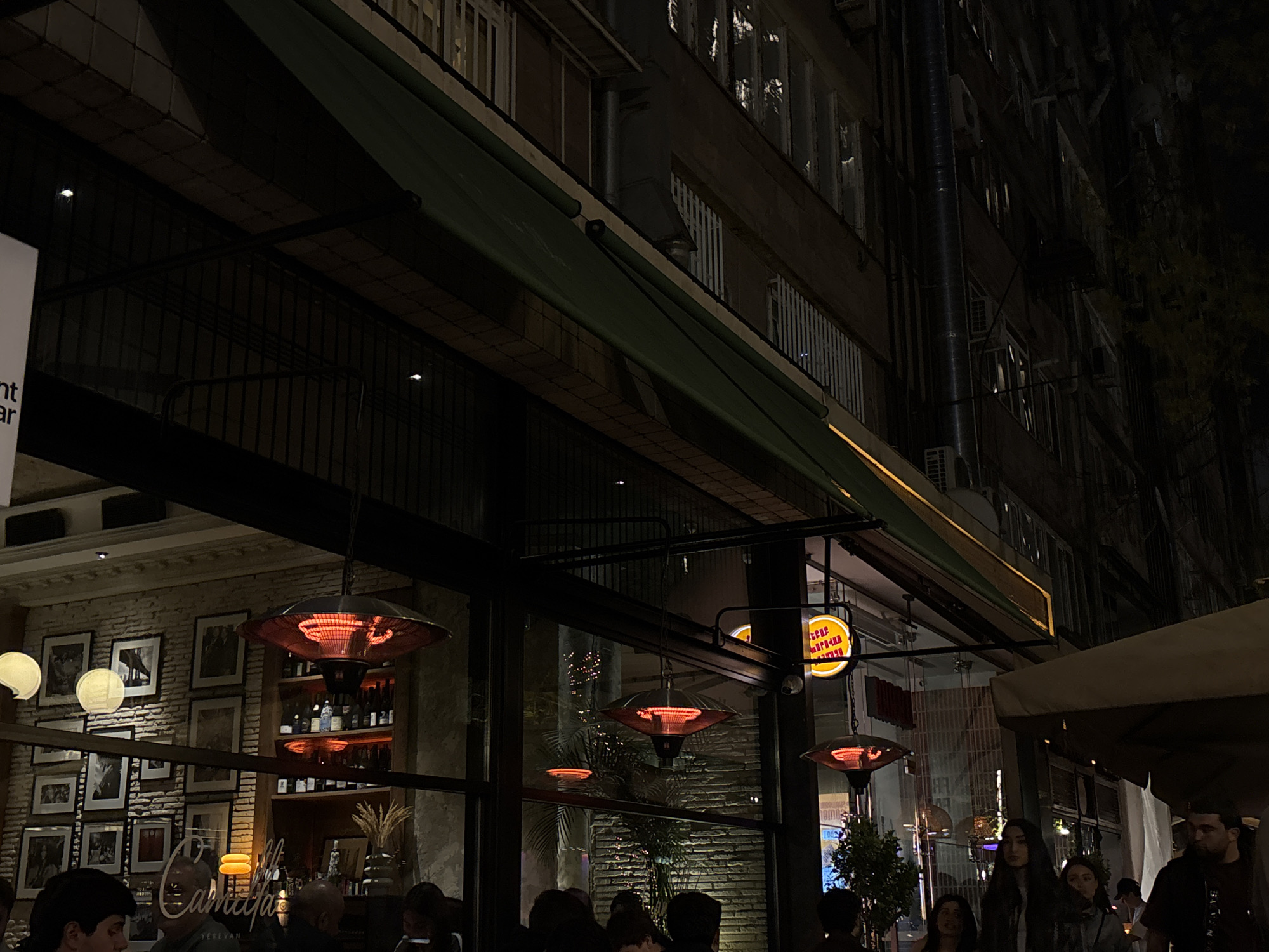 The image depicts a vibrant nighttime scene at a restaurant or cafe in an urban setting, identified as Yerevan, Armenia. The establishment features large glass windows, allowing a view into its warmly lit interior and revealing an outdoor seating area.
In the foreground and middle ground, numerous people are present, both inside the restaurant and in the outdoor space. They are casually dressed and appear to be socializing. Several bright red-glowing outdoor patio heaters are prominently visible, hanging from the ceiling structure, suggesting a cool evening and creating a warm ambiance.
Through the glass, the interior of the restaurant shows exposed brick walls decorated with an array of framed pictures. Shelves stocked with bottles, likely alcoholic beverages, are also visible. Hanging spherical lights illuminate the inside. A sign inside clearly displays the words "Camilla" and "YEREVAN," confirming the location.
Above the restaurant's facade, a long green awning extends. The upper part of the image shows the dark exterior of a multi-story building with numerous windows reflecting light. A large dark pipe runs vertically up the building, and various air conditioning units are visible on its facade. On the right, a yellow circular sign with Armenian text "ԵՐԵՎԱՆ ՊԱՐԿՈՎԵՂ" is illuminated. A partial white sign on the far left reads "nt ar." The overall scene portrays a lively and inviting atmosphere during the evening hours.