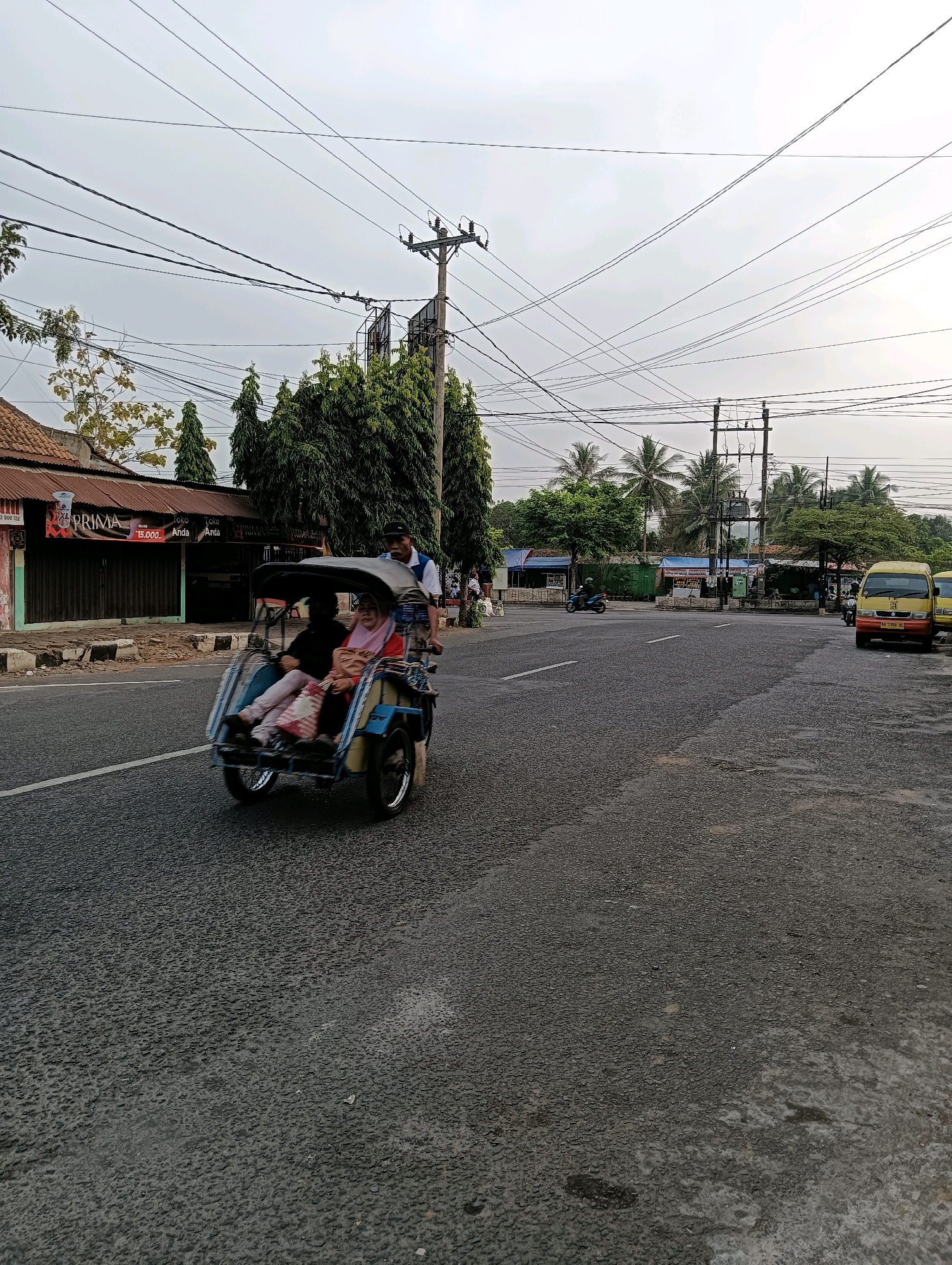 In Purworejo, Indonesia, a pedestrian three-wheeled vehicle, commonly known as a "becak," is traveling on a paved road. The becak is occupied by three individuals: a driver pedaling at the rear and two passengers in the front seating area. The passengers appear to be two women. One woman is wearing a pink headscarf and a patterned dress, while the other, seated behind her, is wearing dark clothing. The becak itself is blue and yellow.
The scene unfolds under an overcast sky, with a backdrop of trees and utility poles supporting numerous electrical wires, suggesting an urban or semi-urban environment. To the left of the road, a building with a corrugated metal roof and storefronts is visible. One storefront displays a sign with the word "PRIMA" in red lettering. Further down the road, to the right, a yellow minibus with the license plate "AA 1393 BG" is parked. A motorcycle is also seen on the road, heading in the opposite direction of the becak. The asphalt road surface shows signs of wear and some wet patches. The overall impression is of a typical street scene in an Indonesian city.