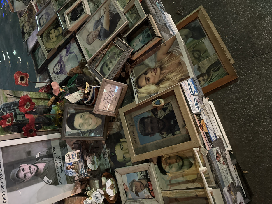 A cluttered memorial displays numerous framed photographs and portraits. Red poppy-like flowers and other mementos are interspersed among the images. Hebrew text appears on various items, possibly names, dates, or tributes. The photographs feature both men and women, some in military uniforms and others in casual attire. The arrangement of the photographs creates a layered, dense collage of remembrance.