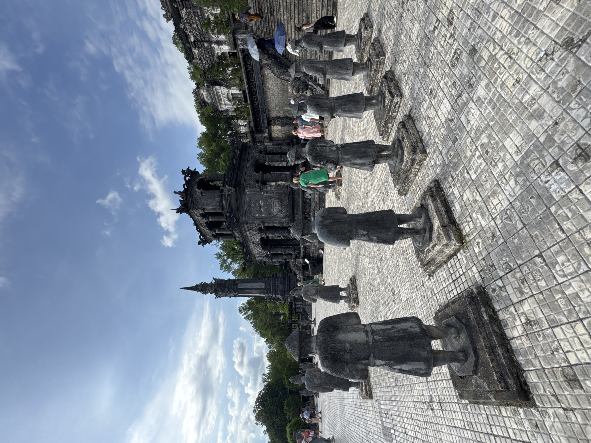 The image depicts the exterior of a historical complex, likely a tomb or temple, located in Thủy Xuân Ward, Vietnam. In the foreground, a paved courtyard is lined with several life-sized stone statues of figures wearing traditional attire and conical hats. These statues are arranged in rows, facing away from the viewer, towards a large, dark, multi-tiered structure in the background.
This main structure is elaborate, featuring intricate carvings, arched doorways, and multiple levels of ornate roofs. To the left of this structure, a tall, slender, spire-like monument rises towards a partly cloudy blue sky. To the right, a grand staircase with stone balustrades ascends towards more buildings and forested hills.
Several people are scattered throughout the scene. Some are in the mid-ground, walking amongst the statues, while others are near the main structure, ascending the stairs or observing the architecture. Some individuals are holding umbrellas, suggesting it might be sunny with a chance of rain. The overall atmosphere suggests a historical tourist site, with elements of architecture, sculpture, and human activity under a bright, daytime sky.