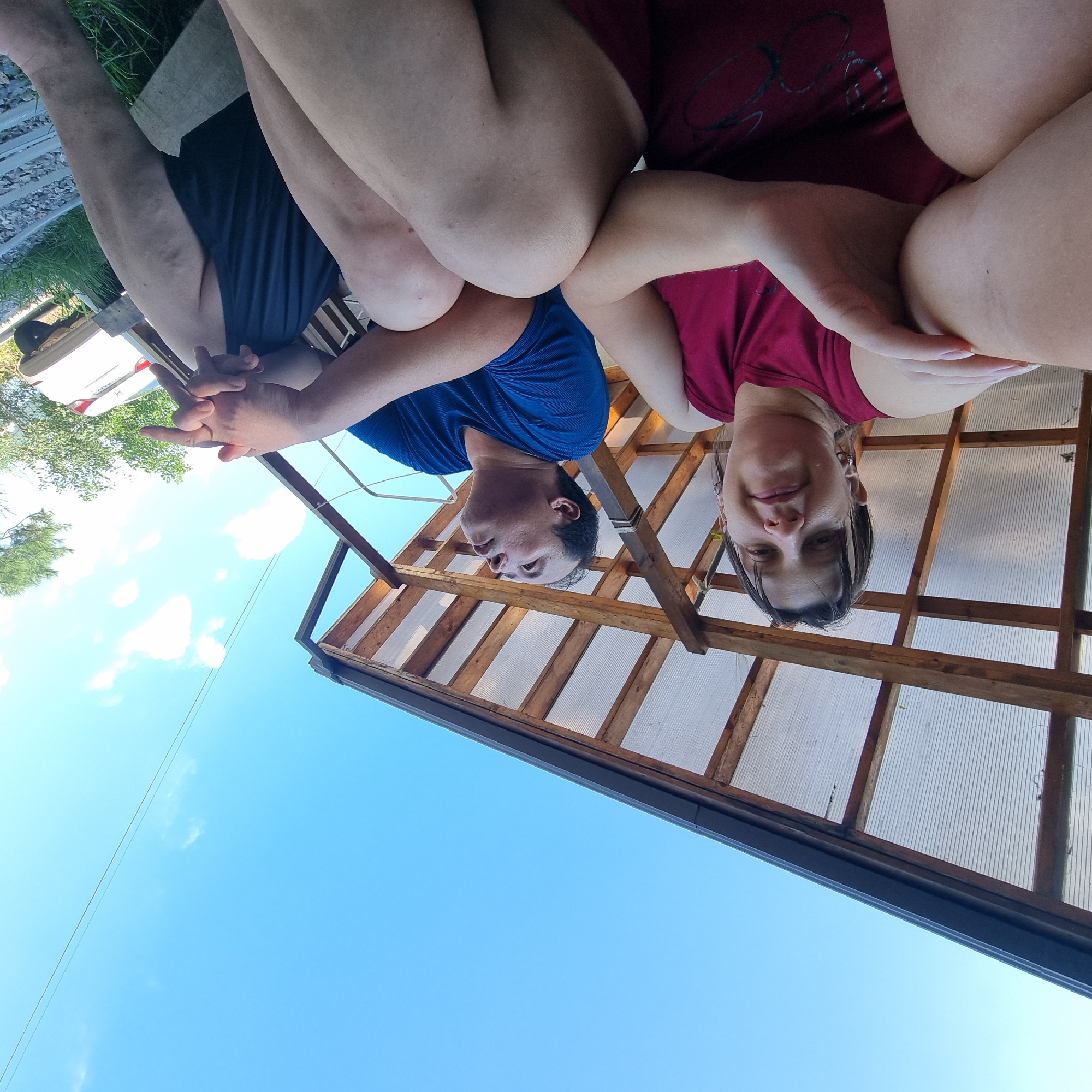 This is a ground-level, wide-angle, eye-level photograph taken outdoors on a clear, sunny day in Dunnae-myeon, South Korea. The image features two individuals, a woman and a man, seated side-by-side under a wooden structure with a translucent roof. The woman, on the left, is wearing a maroon tank top and looking towards the camera with a slight smile. Her left arm is resting on her lap, and her right arm is bent across her chest. The man, on the right, is wearing a blue t-shirt and dark shorts. He is looking upwards and to the right, with his hands clasped in his lap. In the background, a white car is visible, along with trees and a power line stretching across the bright blue sky. The ground appears to be covered with gravel and grass.