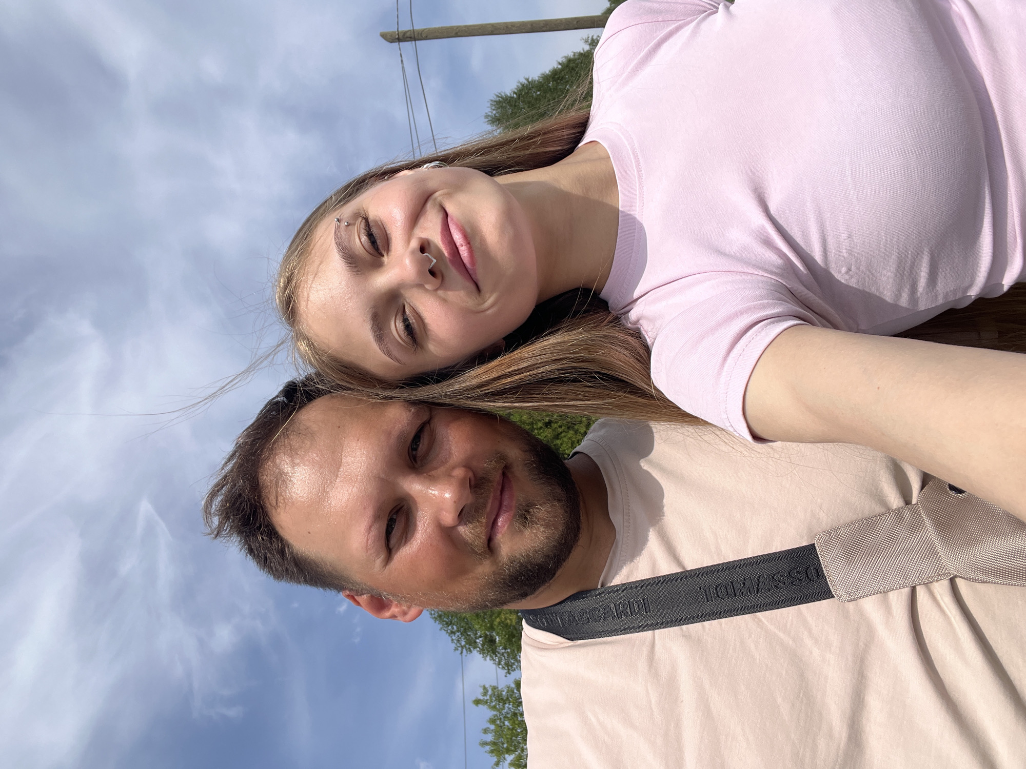 A selfie captures a man and a woman lying side-by-side outdoors, likely in a park or natural setting. The woman, positioned above the man, has fair skin, light brown hair, and a piercing in her eyebrow and nose. She wears a pink t-shirt and smiles at the camera. Below her, the man has dark hair and a beard. He wears a beige t-shirt with a black shoulder strap bearing the name "BORLMARSSDI". The background reveals a blue sky with scattered clouds, trees, and a power line. There is nothing to suggest the image was taken in Фоки, Russia.