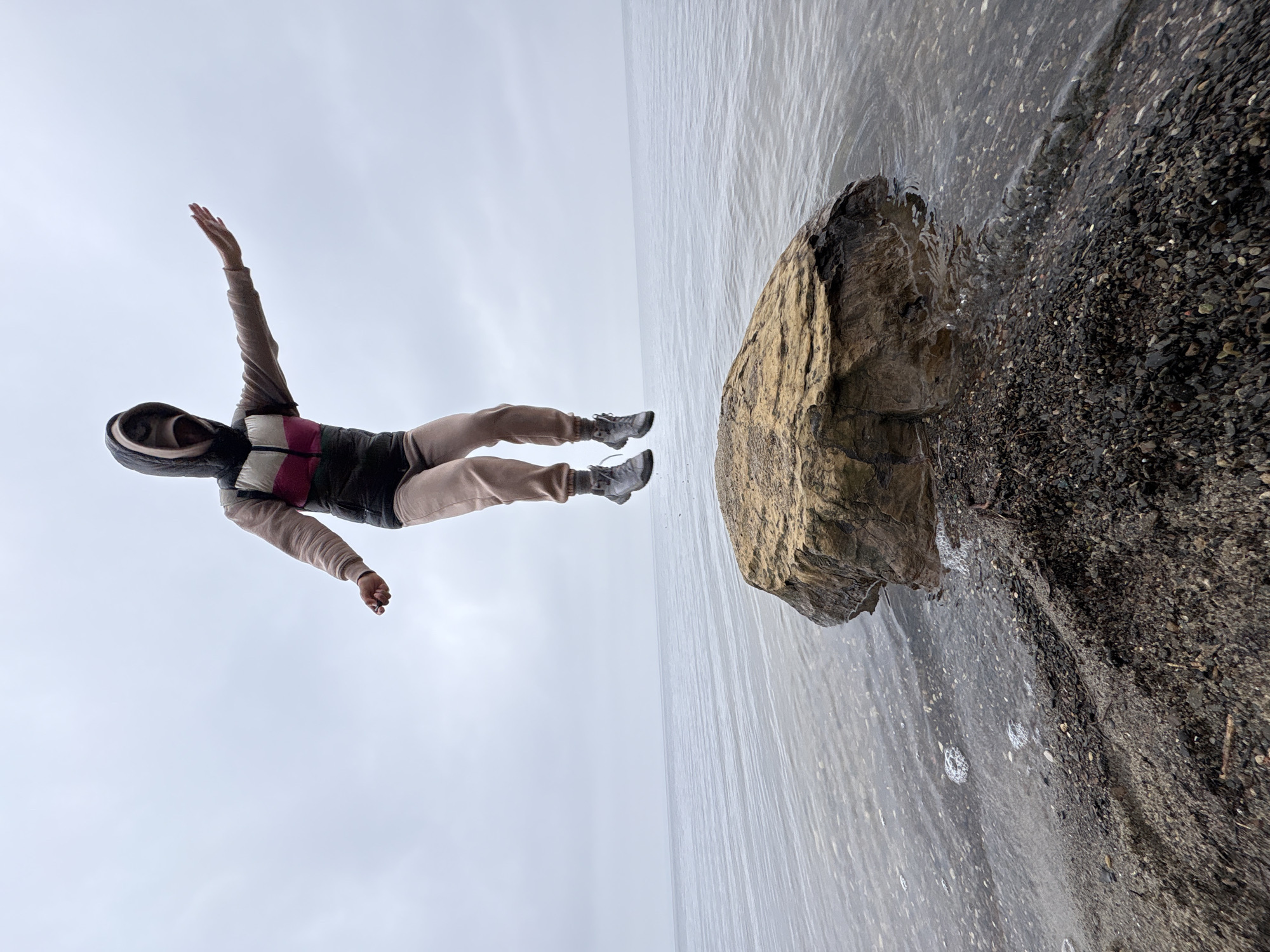 A person wearing a hooded jacket, light-colored pants, and sneakers is airborne next to a body of water and a rocky shore. The person has outstretched arms. The location appears to be on the shoreline of the Volga River, near Ulyanovsk, Russia. The sky is overcast. A large rock is partially submerged in the water. The shoreline consists of small pebbles and dark-colored soil.