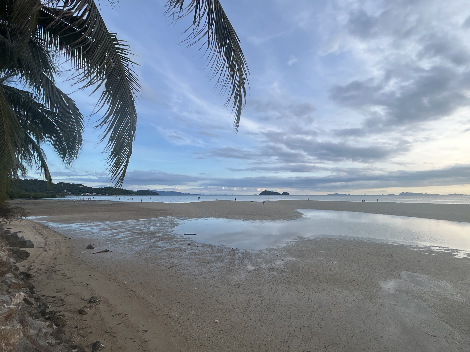 The image depicts a beach scene in ตำบลเกาะพะงัน, Thailand. In the foreground, there's a sandy beach transitioning into a shallow, reflective body of water, likely a tidal pool. The sand appears damp and textured. Several people are walking along the beach in the midground. In the background, there are islands or land masses visible across the water, with a cloudy sky above. Palm tree fronds hang into the frame on the left side, partially obscuring the view.