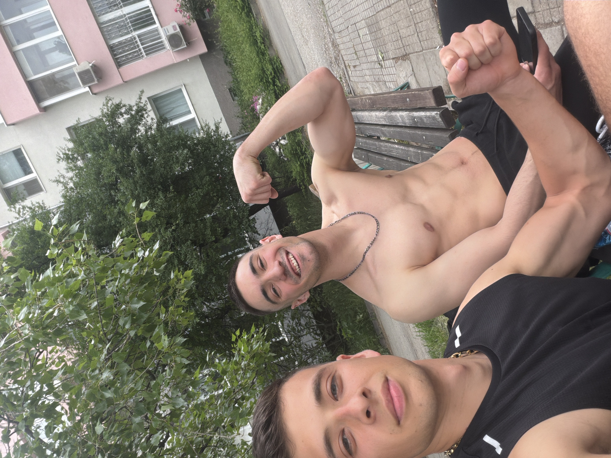 The image captures two men in an outdoor setting. The shirtless man in the background, wearing only black shorts and a silver chain, sits on a wooden park bench with his right arm raised, flexing his bicep. He is smiling and has a dark complexion. The man in the foreground is wearing a black tank top with white stripes, as well as a gold chain and is looking at the camera. The location appears to be a public park with a building in the background featuring pink and white exterior paint. There is a green tree with leafy branches beside them, as well as paved stone around the area. Considering the context, it's reasonable to believe this scene occurs in Plovdiv, Bulgaria.