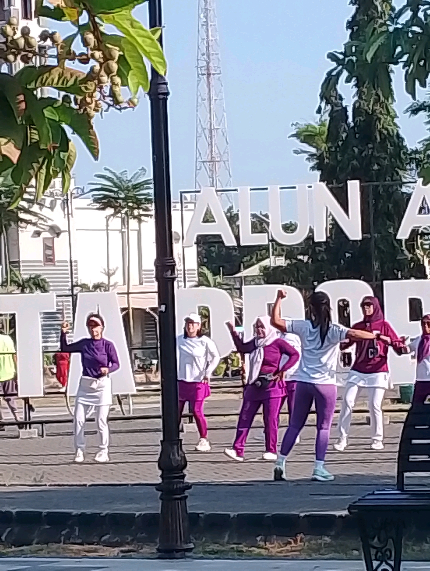 In Probolinggo, Indonesia, a group of women are engaged in outdoor exercise. The scene is set in a public space, likely a town square or park, with large white lettering in the background that reads "ALUN A" and "TA". This suggests the location is Alun-Alun, a common public square in Indonesian cities, and this particular one is in Probolinggo. The women are dressed in athletic wear, with many wearing headscarves and long pants or skirts. They appear to be participating in a synchronized aerobics or dance routine, with some individuals striking poses and others performing arm movements. The environment is sunny, with a clear blue sky, and trees and buildings are visible in the background. A lamppost stands in the foreground, and a park bench is partially visible on the right side of the image. The overall atmosphere is one of healthy activity and community engagement.