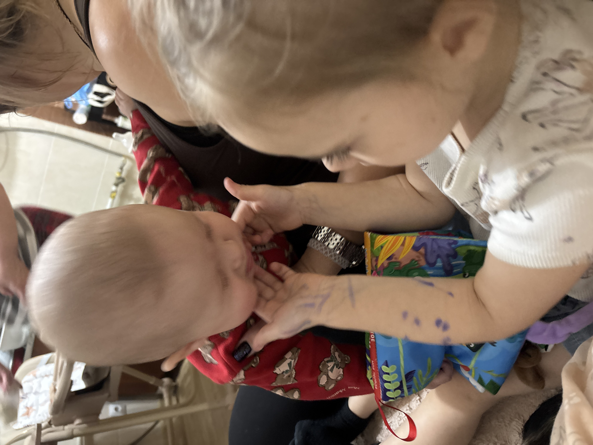 A close-up, high-angle shot depicts a baby, wearing a red patterned top, being held by an adult. A child with blonde hair is leaning close to the baby, interacting with them. The child has what appear to be blue pen marks on their arm. There are other adults in the background, partially visible. The location is unspecified; there is no information in the image that supports the presence of Sochi, Russia. The environment includes medical equipment and what appear to be a soft book.