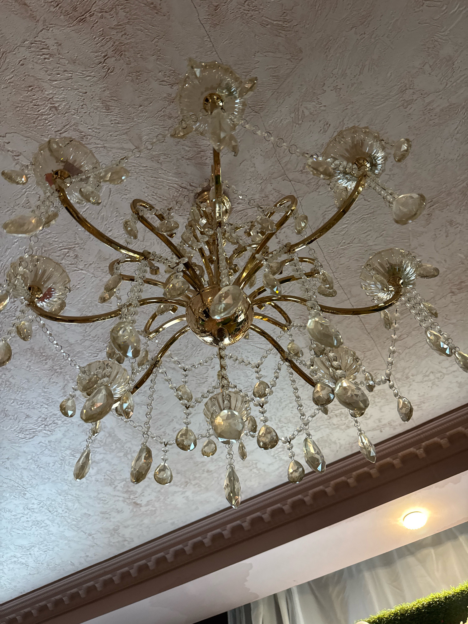 A crystal chandelier with a gold-colored frame hangs from a textured white ceiling. The chandelier has multiple arms, each adorned with clear, faceted crystal drops and decorative crystal pieces resembling flowers. The crystals catch the light, creating a sparkling effect. The ceiling has a textured pattern. A dark brown decorative molding runs along the edge of the ceiling. A bright light fixture is visible on the ceiling in the lower right portion of the image, and a glimpse of curtains and greenery can be seen in the lower part of the frame. The image appears to be taken from below, looking upwards at the chandelier. This scene is not in Torzhok, Russia.