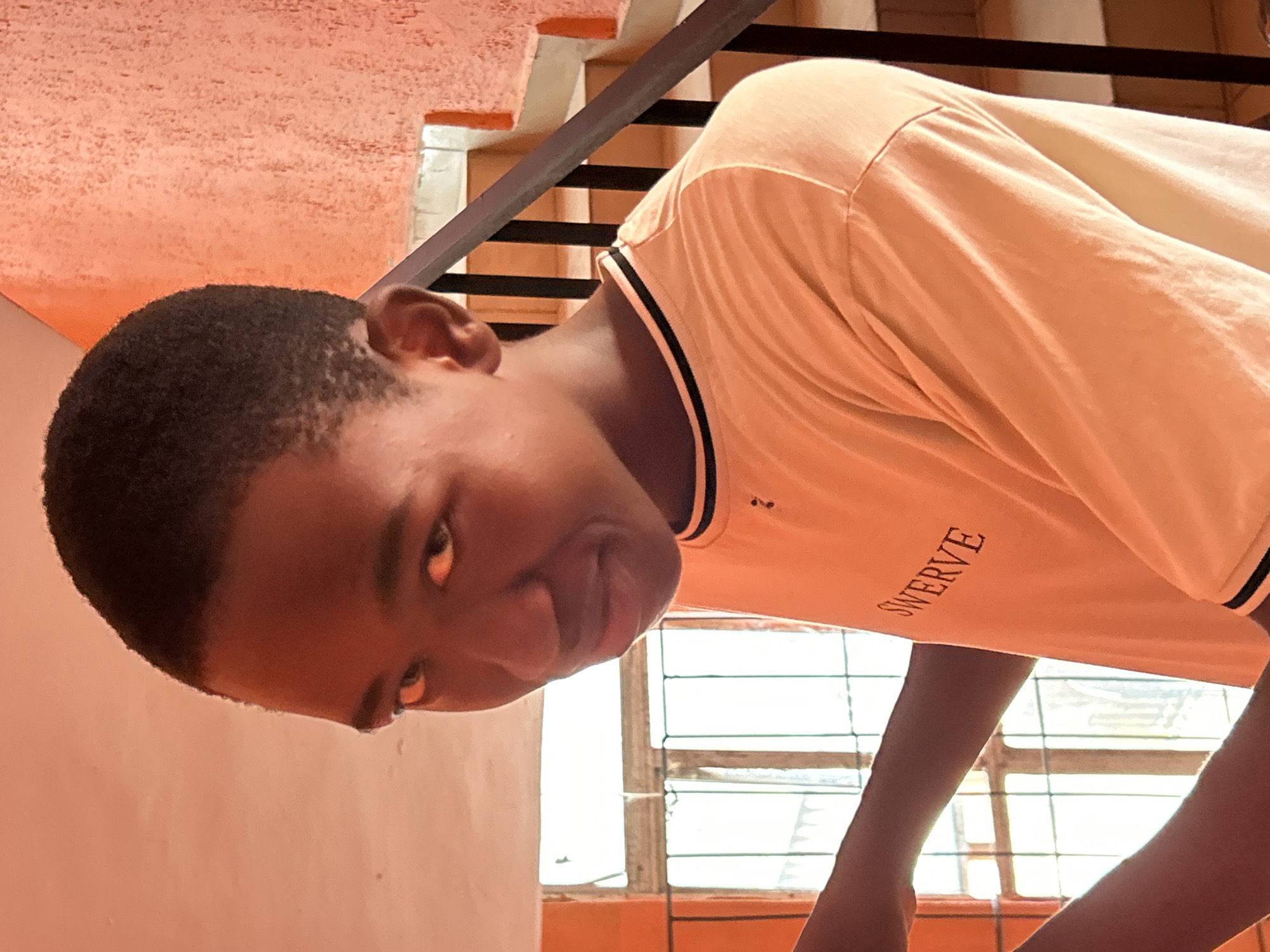 A young, dark-skinned man with short, cropped hair is seen indoors, possibly in a residence in Ihiagwa, Nigeria. He is wearing a light orange t-shirt with a black and white striped collar and the word "SWERVE" printed on it. He appears to be either bending or looking down at something out of frame. A metal staircase and a barred window are visible in the background. The walls are painted a peach color.