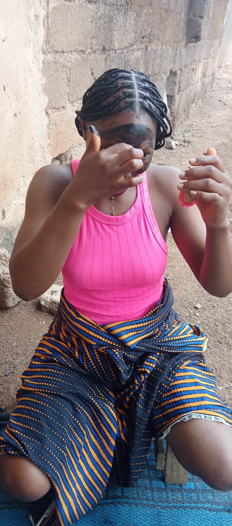 A woman is sitting outdoors in Akwanga, Nigeria. She has braided hair and is wearing a pink tank top and a dark blue and yellow patterned wrap-around skirt. She is applying a dark substance to her face, possibly makeup or traditional paint. She is holding a small container in her right hand. The environment consists of a simple dirt ground, a blue woven mat, and a wall made of mud or brick in the background.