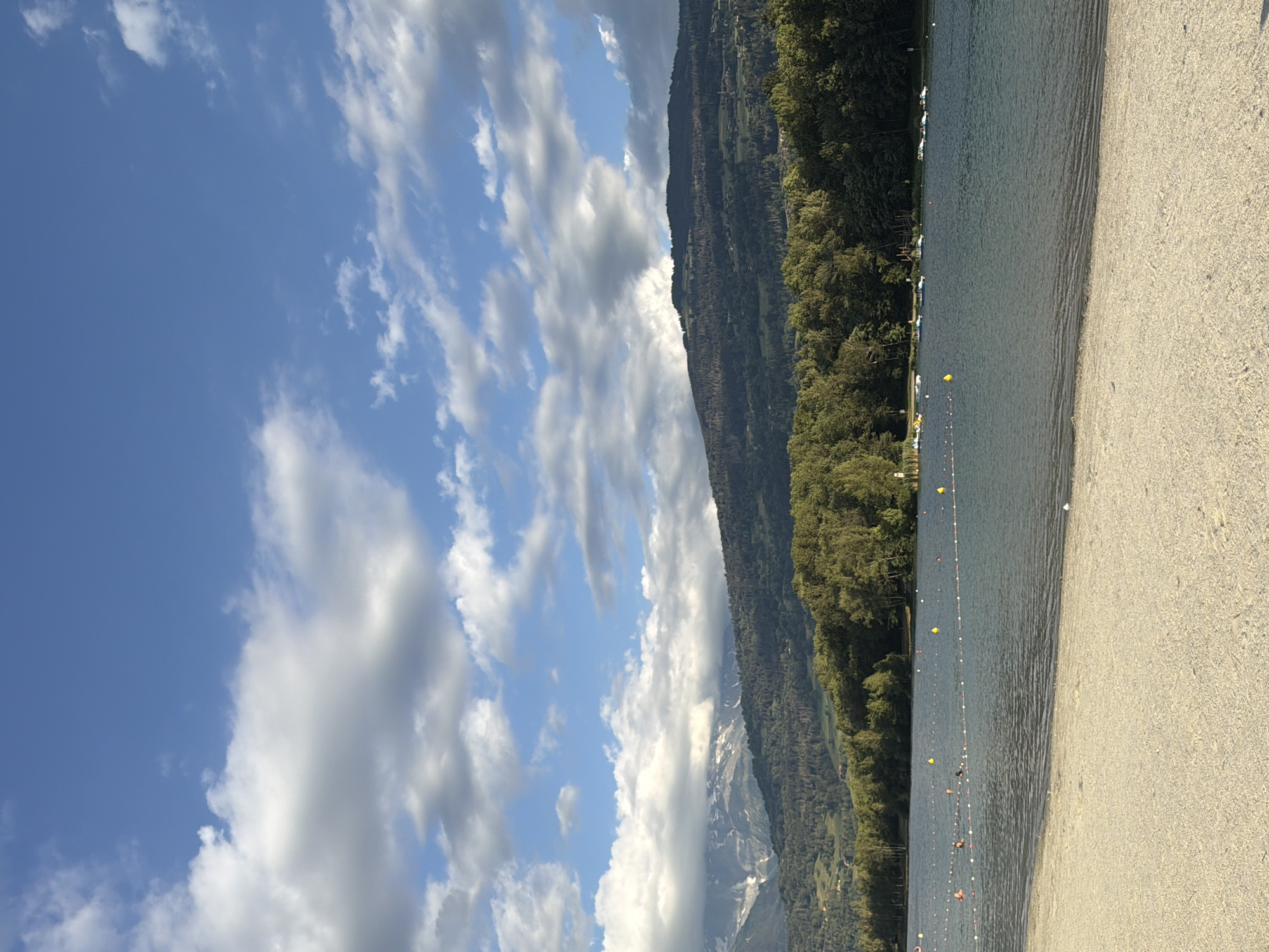 The image depicts a scenic view of Lake Passy in Passy, France, on a clear day with scattered clouds. The foreground features a sandy beach, leading to the calm waters of the lake. A roped-off swimming area is visible in the lake, marked by buoys and floats. Several people are in the water, with some swimming and others wading near the shore. A line of small boats, including pedal boats and a sailboat, are moored along the far bank of the lake. Beyond the lake, a dense forest covers the lower slopes of a mountain range. The mountains are partially shrouded in clouds, with some snow-capped peaks visible in the distance. The sky is a vibrant blue, with white and gray clouds scattered across it. The overall impression is one of a peaceful and idyllic summer day at a popular tourist destination.