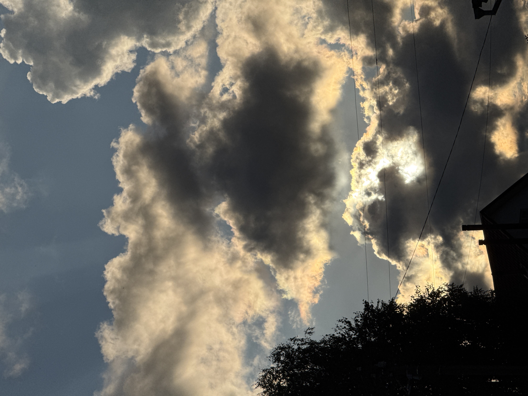 This is a medium shot, eye-level, outdoor, daytime photograph taken in Ветлужский, Russia. The image depicts a dramatic sky filled with a mix of dark and light clouds, illuminated by the sun which is partially obscured. The sunlight creates an iridescent effect on some of the clouds, casting a golden and rainbow-like hue. In the lower left quadrant, the dark silhouette of a tree with leaves is visible, its branches reaching towards the sky. Below the tree, the roofline and chimney of a building are silhouetted against the bright sky. Several thin wires, likely utility lines, stretch horizontally across the frame, intersecting the clouds and the building. The overall mood is one of a dynamic and possibly stormy sky, with pockets of intense light breaking through.
