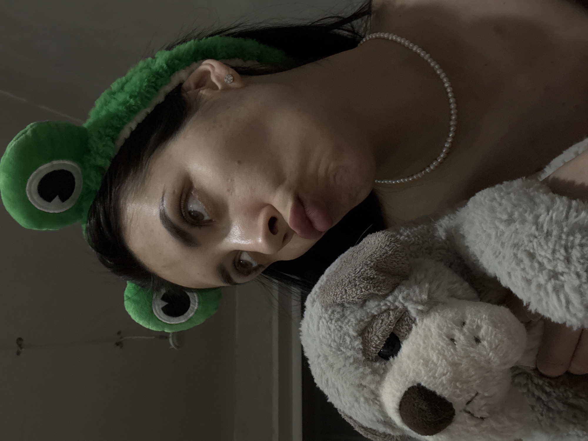 A person is depicted in a close-up, angled view, holding a plush, gray and white stuffed animal. The person is wearing a green, frog-themed headband and a pearl necklace. A small earring is visible in the person's ear. The background is dimly lit and suggests an indoor setting, potentially a bedroom, due to the casual attire and presence of the plush toy. There is no information to suggest the location is Votkinsk, Russia.