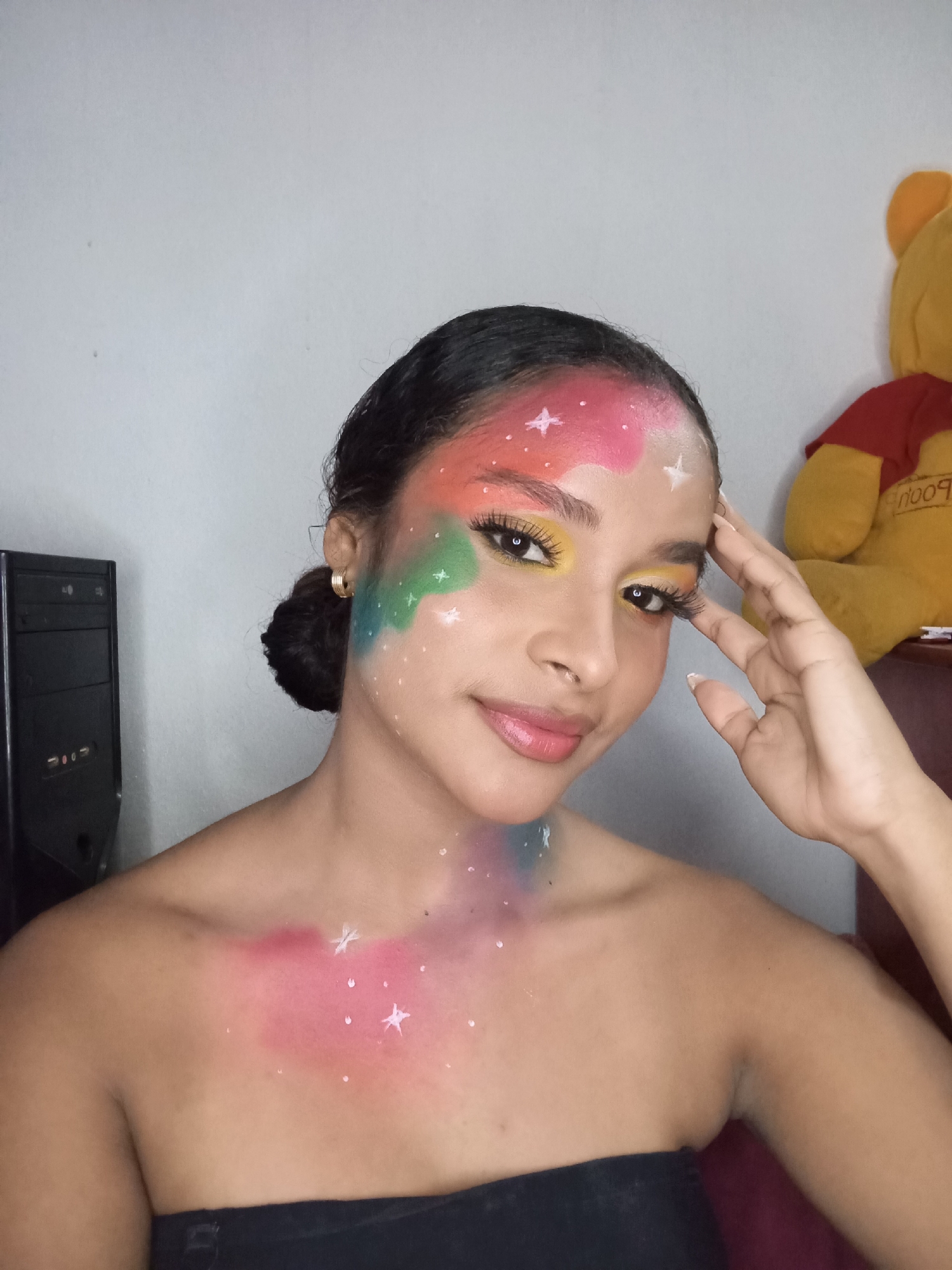 A young woman is posing indoors, adorned with brightly colored makeup that extends from her forehead and face down to her shoulder. The makeup features a nebula-like design with hues of pink, green, and blue, and is sprinkled with white star shapes. She is wearing minimal clothing, likely a black strapless top. Her hair is pulled back into a bun, and she is wearing small gold hoop earrings. Her right hand is raised near her forehead.
Behind her, on the left, is a black computer tower. To her right and slightly behind her is a stuffed Winnie the Pooh toy sitting on a surface. The background wall is a light gray color.
The location, Juan Griego, Venezuela, is not discernable from the image.