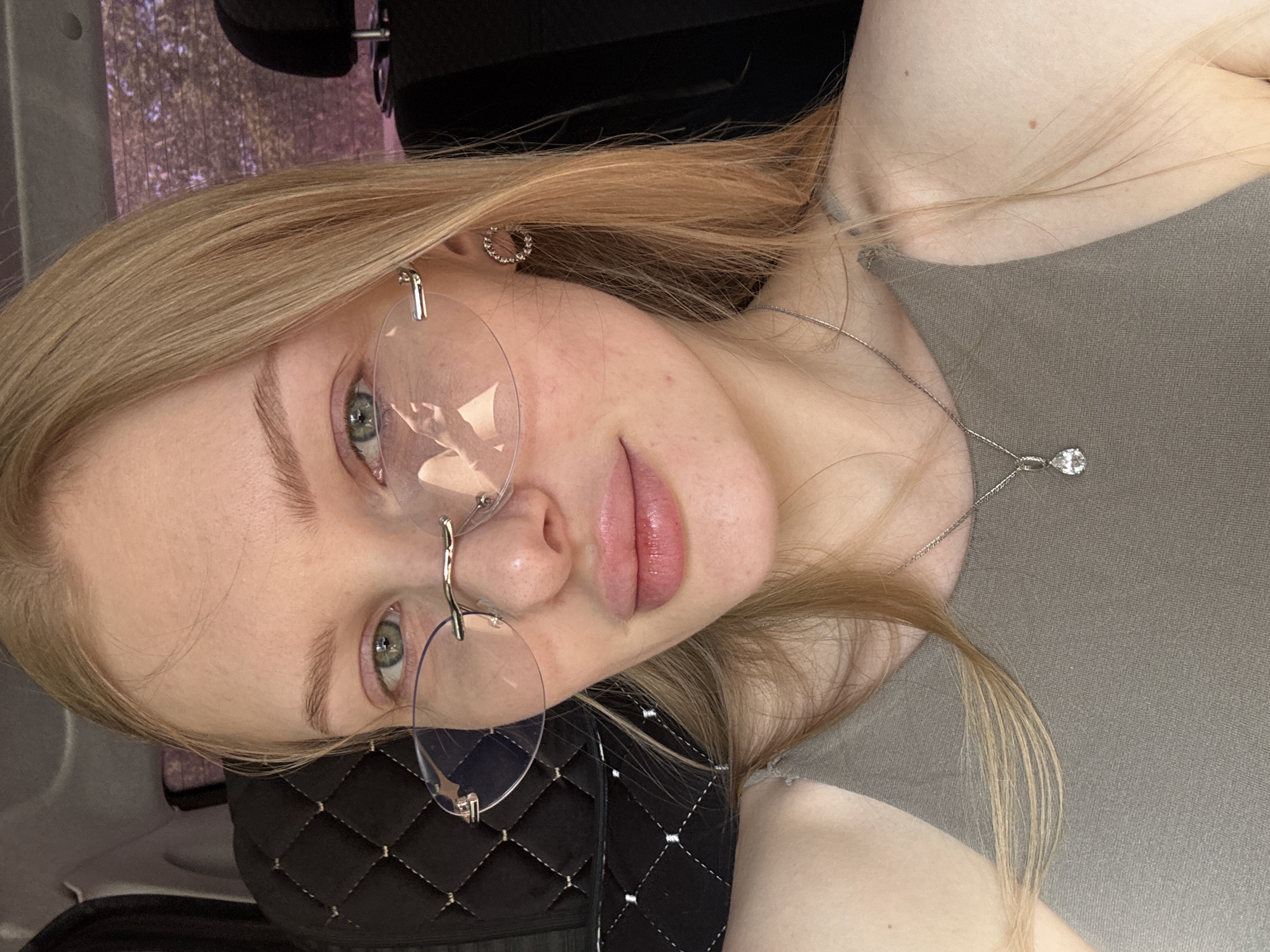 A young woman with blond hair and light skin is shown in a selfie. She is wearing rimless glasses and a silver necklace with a teardrop-shaped pendant. She has an earring in her left ear. She's wearing a gray top. The background appears to be the interior of a car, with black quilted fabric visible. The setting appears to be in Perm, Russia.