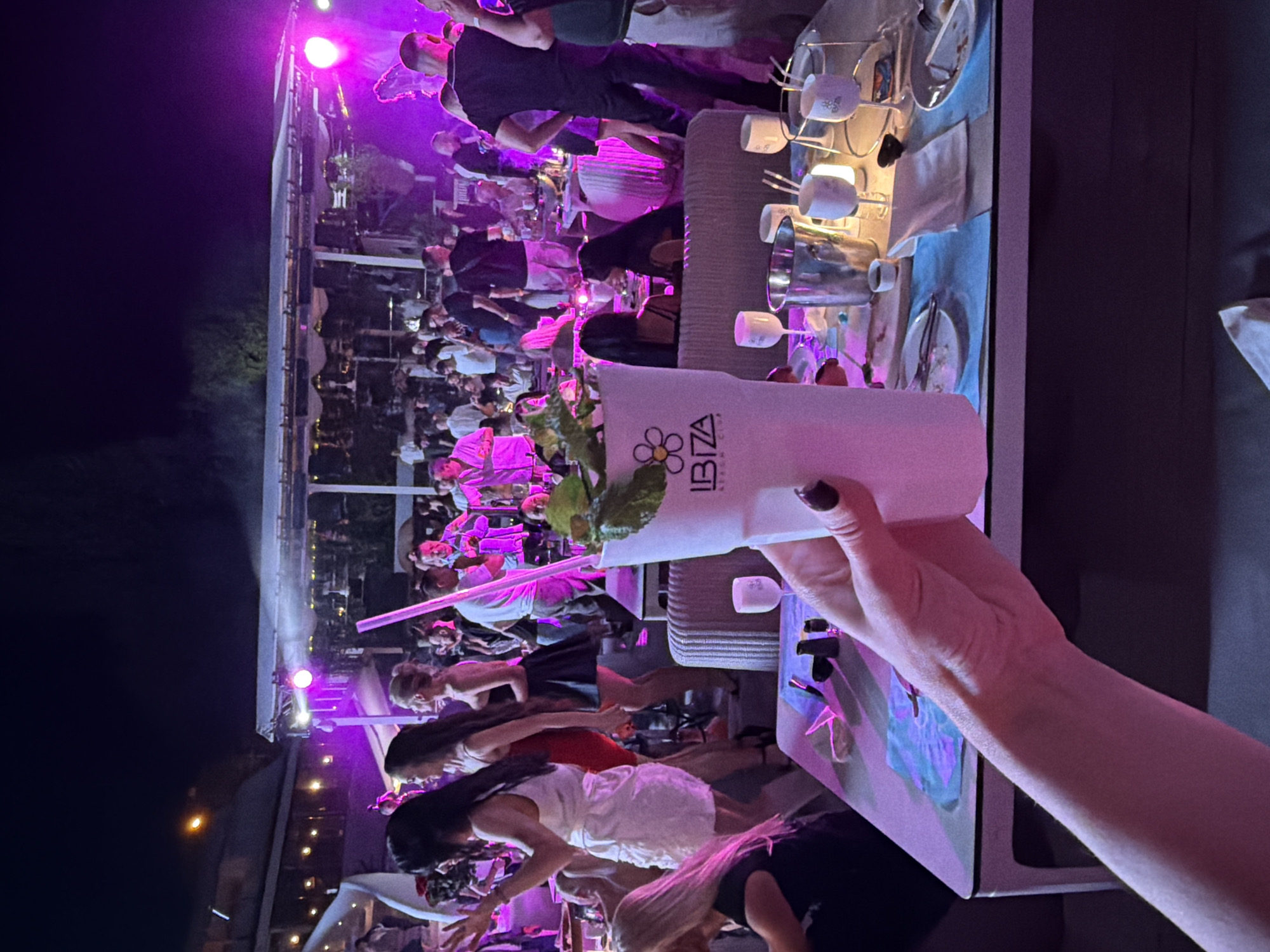 The image shows a nighttime scene at an outdoor venue with purple lighting. In the foreground, a person's hand holds a tall white cup with a green garnish and a pink straw. The cup has the "Ibiza Beach Club" logo on it. The background depicts a crowded gathering of people, some of whom are dancing or standing around tables. A stage with lights is visible in the distance, suggesting a performance or event is taking place. The presence of the "Ibiza Beach Club" logo indicates the location is likely a beach club. While the image itself doesn't explicitly state the city, if the venue is "Ibiza Beach Club" in Odesa, Ukraine, then the scene is set there. The overall atmosphere appears festive and energetic, with people dressed in casual and semi-formal attire, indicative of a night out.