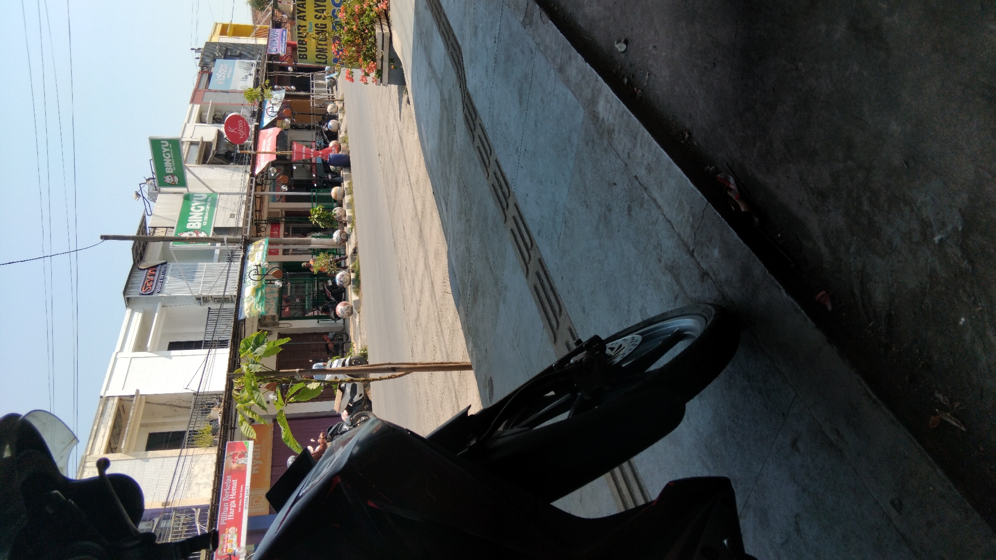 The image captures a street scene in Batang, Indonesia, likely on a sunny day given the clear sky and strong shadows. In the foreground, the lower portion of a black motorcycle is visible, with its front wheel and part of the frame occupying the left side of the frame. The motorcycle appears to be parked or stationary on a paved sidewalk. The sidewalk itself is made of concrete and features raised tactile paving for visually impaired pedestrians.
Beyond the sidewalk, a street runs horizontally across the image. Several motorcycles are parked along the side of the street, indicating a commercial area. On the opposite side of the street, a row of multi-story buildings lines the road. These buildings appear to house various businesses, as evidenced by the signage visible. Signs such as "SAMN," "BINGYU," and "LaSalon" are legible, suggesting the presence of shops, cafes, or service establishments. Some of these buildings have awnings providing shade over the storefronts.
The environment is urban, with buildings and paved surfaces. There are some potted plants and decorative spherical bollards along the street, adding a touch of landscaping. A few individuals are discernible in the background, near the storefronts and on the street, but their activities are not clearly defined due to the distance and image resolution. The overall impression is that of a bustling commercial street with everyday urban activity.