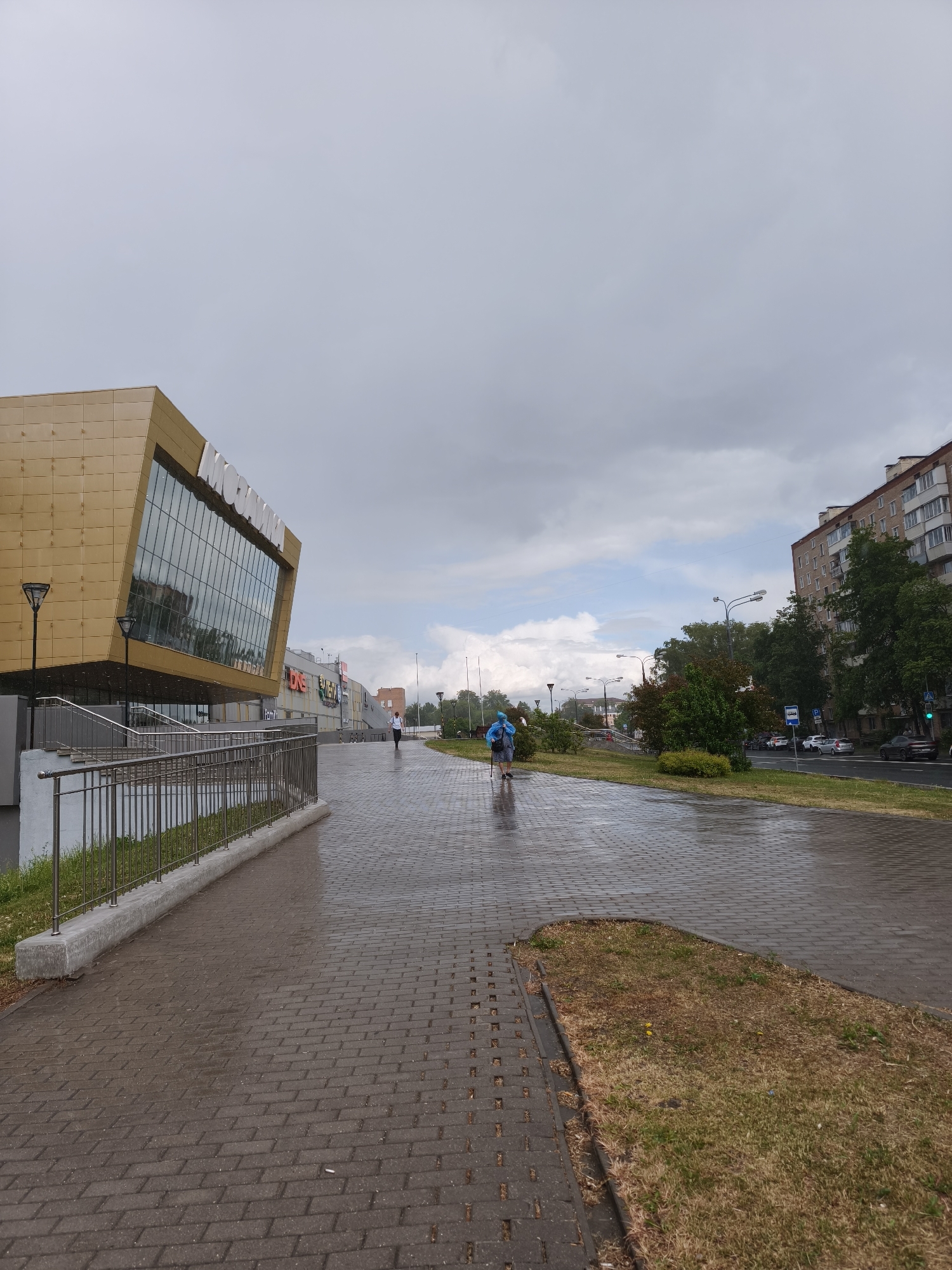 The image depicts a street scene in Moscow, Russia on a wet day. The ground is paved with wet cobblestones, reflecting the overcast sky. A modern building with a gold-toned exterior and large glass windows stands on the left. Multiple storefronts are visible further down the street. A pedestrian is walking on the sidewalk. Across the street, a multi-story residential building can be seen. There are trees and street lights along the roadway. Cars are moving in the distance, adhering to traffic signals.