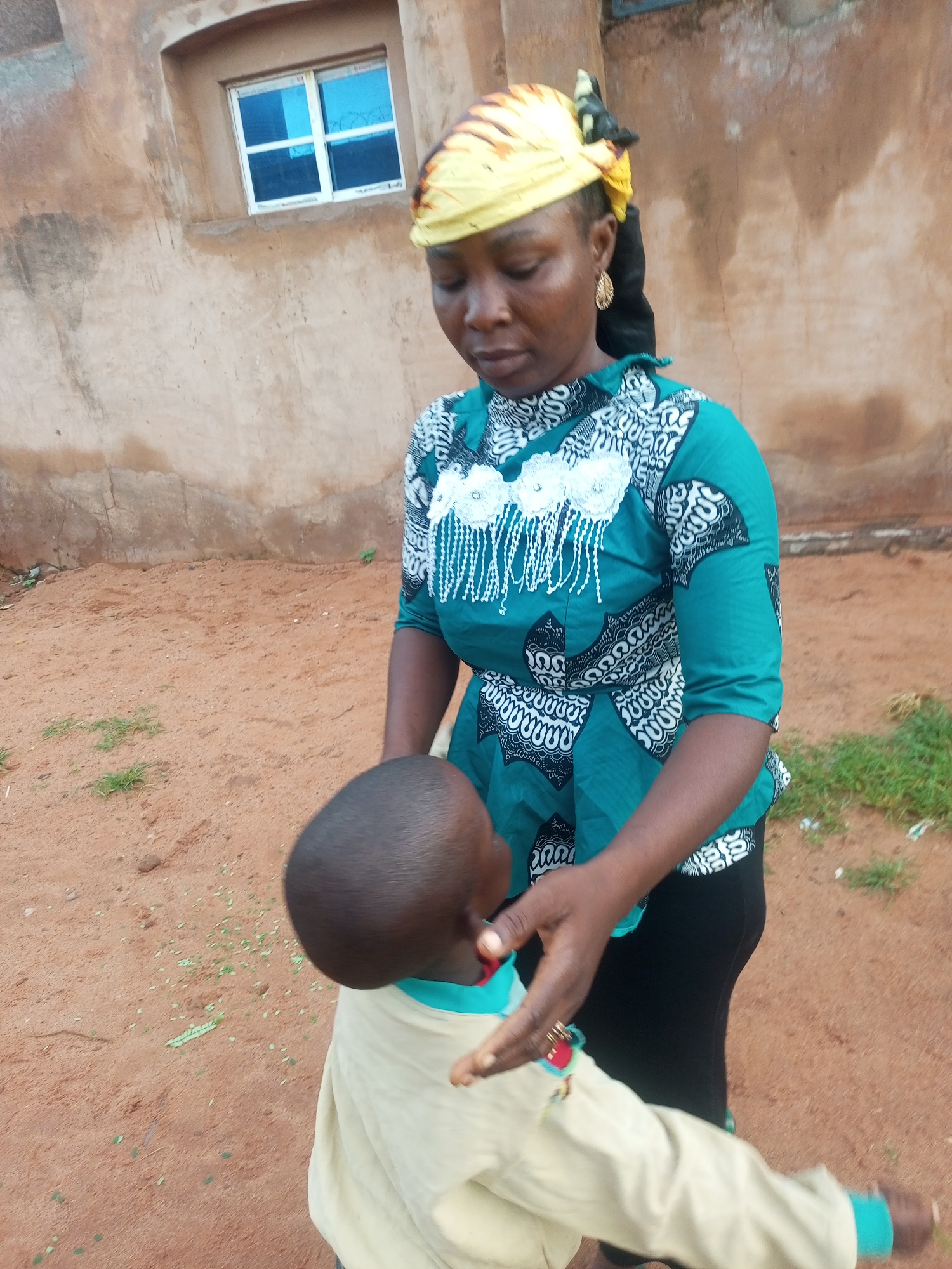 In the city of Tempure, Nigeria, a woman with dark skin is standing outside near a tan, stucco building with a small, blue-tinted window. She is wearing a yellow headwrap patterned with brown and black stripes, a teal and white patterned top with fringe detailing on the chest, and black leggings. She is looking down and holding the arm of a young child, who is wearing a light-colored garment. The ground is a reddish-brown dirt with sparse patches of green grass. The scene appears to be outdoors in a residential area.