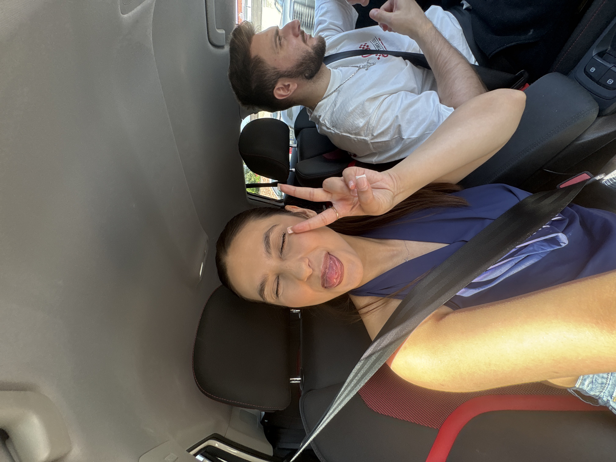 Two people are in a car. A woman in a blue shirt makes a peace sign, sticking out her tongue. The man in the passenger seat wears a white shirt with a chain around his neck, with a black jacket draped over the seat. The interior of the car is grey with red trim on the seats. The exact location in Mönchengladbach, Germany cannot be determined from the image.