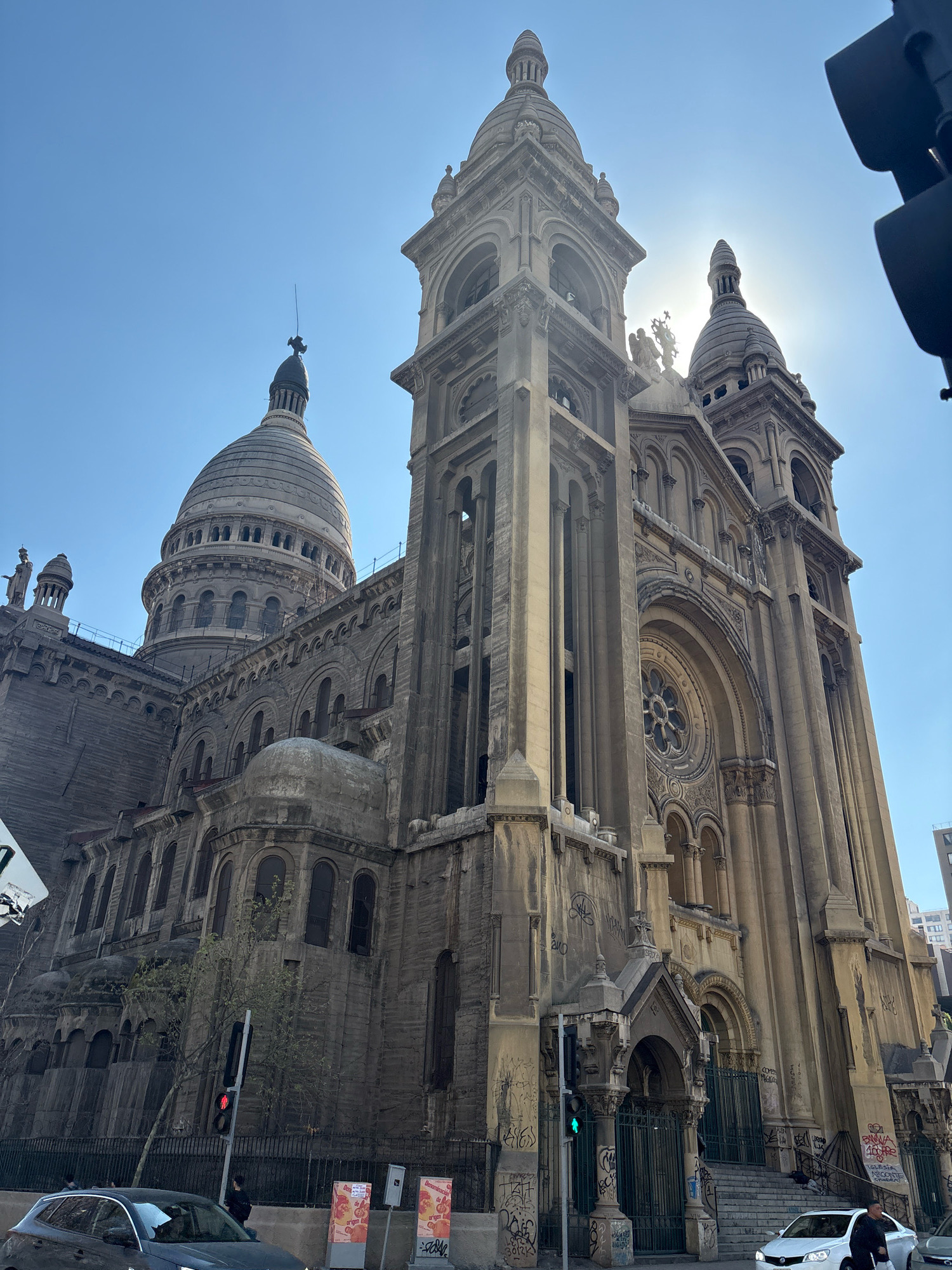 The photograph captures a low-angle, wide shot of the Metropolitan Cathedral of Santiago, Chile, bathed in bright sunlight. The iconic facade of the cathedral dominates the frame, showcasing its intricate architectural details and imposing presence against a clear blue sky. The sun is positioned behind the cathedral's spires, creating a lens flare effect that partially obscures the upper structures.
The cathedral's facade is made of light-colored stone, appearing weathered and showing signs of graffiti in several areas. The building features multiple towers, domes, and arched windows, typical of religious architecture. A large, ornate dome is visible on the left side of the image, while a taller, more slender tower with a spire stands prominently in the center. Smaller spires and decorative elements adorn the rooftops.
In the foreground, a street scene unfolds. A dark-colored SUV is parked on the left side of the road, partially visible. Across the street, a black fence separates the sidewalk from the cathedral's grounds. A traffic light with a red signal is visible, and a person is standing near it. Further down the street, a white sedan is seen. Two red and white advertising stands are also present on the sidewalk. Graffiti is visible on the fence and on some parts of the cathedral's base. The environment appears urban, with a clear sky suggesting daytime.