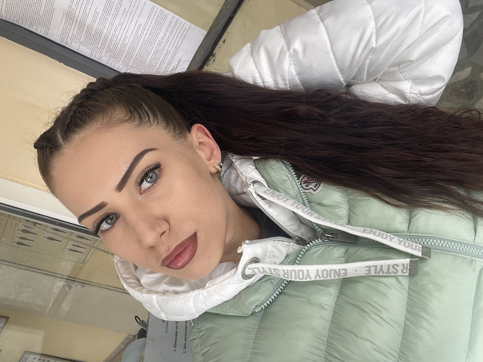 The image depicts a woman with long, dark brown hair styled in a high ponytail with braided sections near the forehead. She has fair skin, defined eyebrows, and striking light blue eyes. She is wearing a light green quilted puffer jacket with a white hood. The zipper pulls of the jacket have "ENJOY YOUR STYLE" printed on them.
In the background, there is a document with text in a Cyrillic alphabet and a glass or countertop surface reflecting some images. A portion of a white quilted surface is also visible in the background, which appears to be part of the jacket's hood.
The woman appears to be taking a selfie or posing for a photograph, looking directly at the camera.
The location of Veliko Tarnovo, Bulgaria, is not directly discernible from the visual information provided in the image.