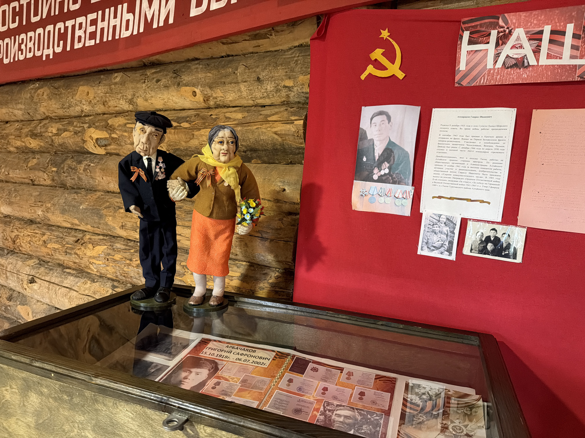 The image depicts a museum display case containing a collection of historical documents and photographs. Two life-sized mannequins of an elderly couple, dressed in period clothing and adorned with medals, stand in front of a wooden wall. The male mannequin wears a dark suit and a black cap, while the female mannequin is in a brown jacket and an orange skirt, holding a bouquet of flowers. Behind the mannequins, a red banner with Soviet imagery, including a hammer and sickle, is displayed. Several framed documents and photographs are pinned to the red banner. The display case in the foreground showcases open books with more documents, photographs, and medals. The overall scene appears to be an exhibit dedicated to commemorating individuals and events from Soviet history, possibly in the city of Ust'-Pyzas, Russia, given the context of the provided information.